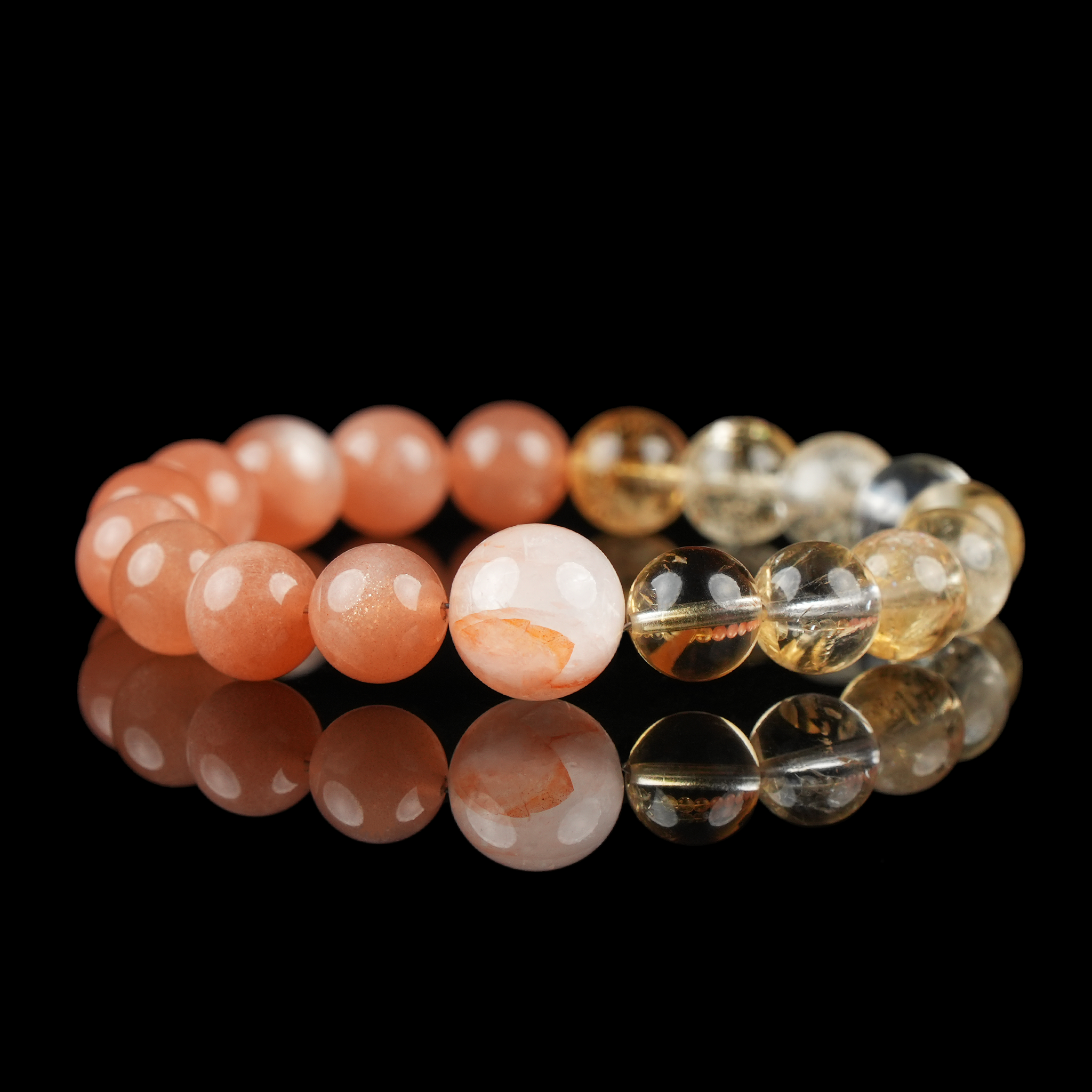 This image showcases a beautifully arranged bracelet composed of polished round beads in warm orange and golden tones. The stones include vibrant orange moonstone, translucent citrine, and red rutilated quartz with fine inclusions. The bracelet is laid flat against a glossy black surface, enhancing the colors and shine of the gemstones.