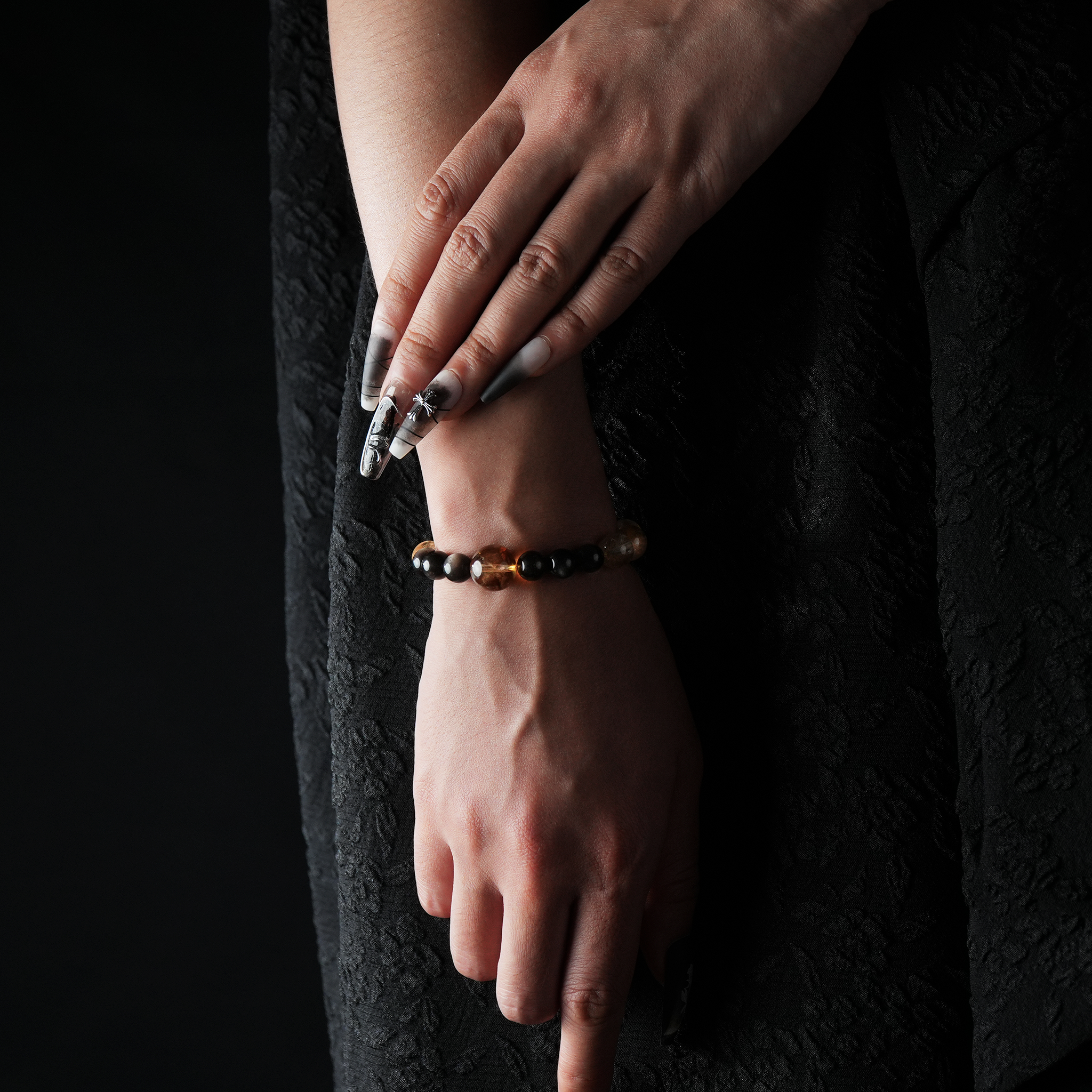 Elegant wrist shot featuring a beaded bracelet composed of citrine, yellow quartz, and silver obsidian gemstones. The woman's hand rests gently against a black patterned dress, with long, detailed nails completing the refined look.