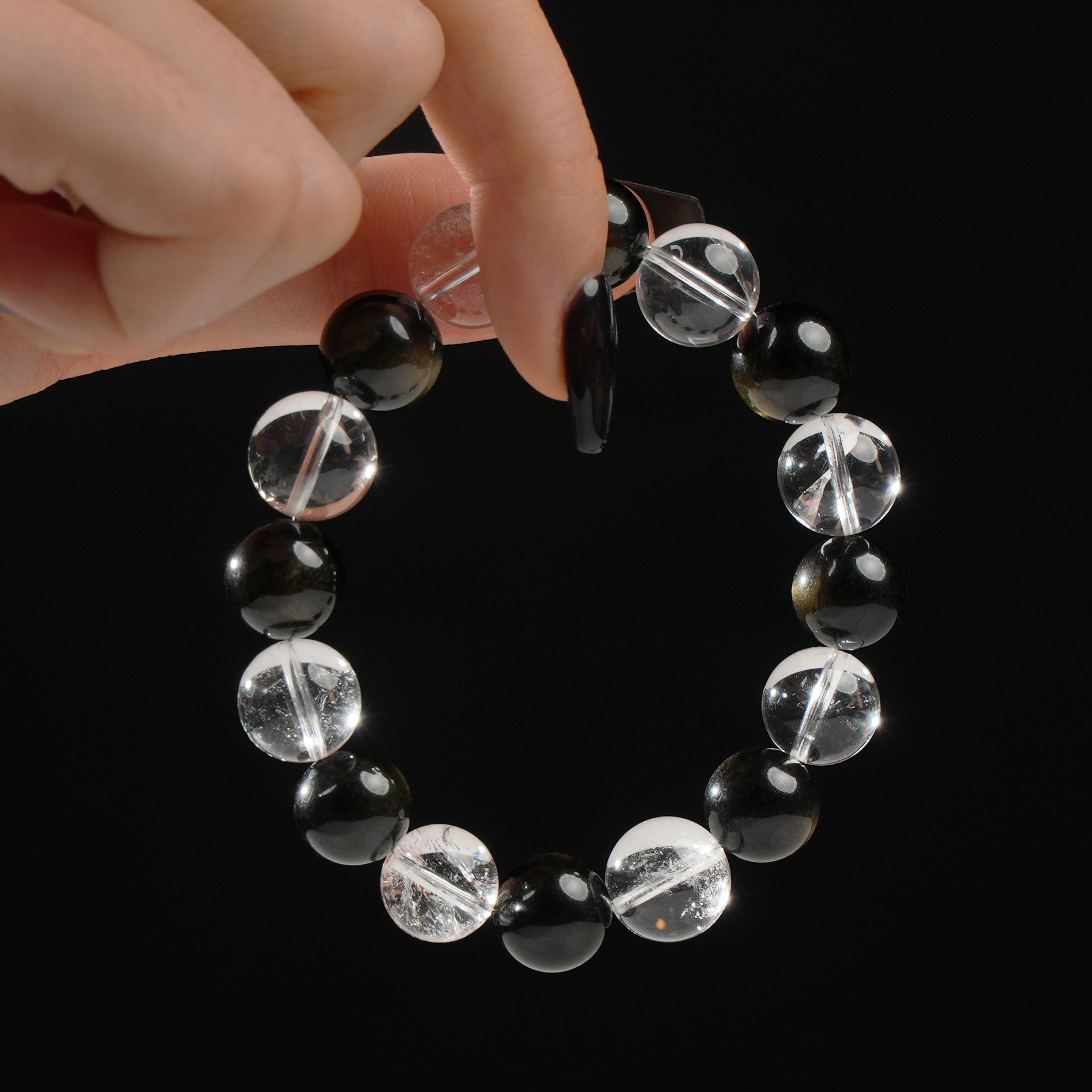 The image shows a hand holding a beaded bracelet composed of 12mm round Clear Quartz and Gold Sheen Obsidian gemstones. The Clear Quartz beads sparkle with clarity while the Gold Sheen Obsidian beads reflect subtle golden highlights, creating a striking contrast against the dark background.