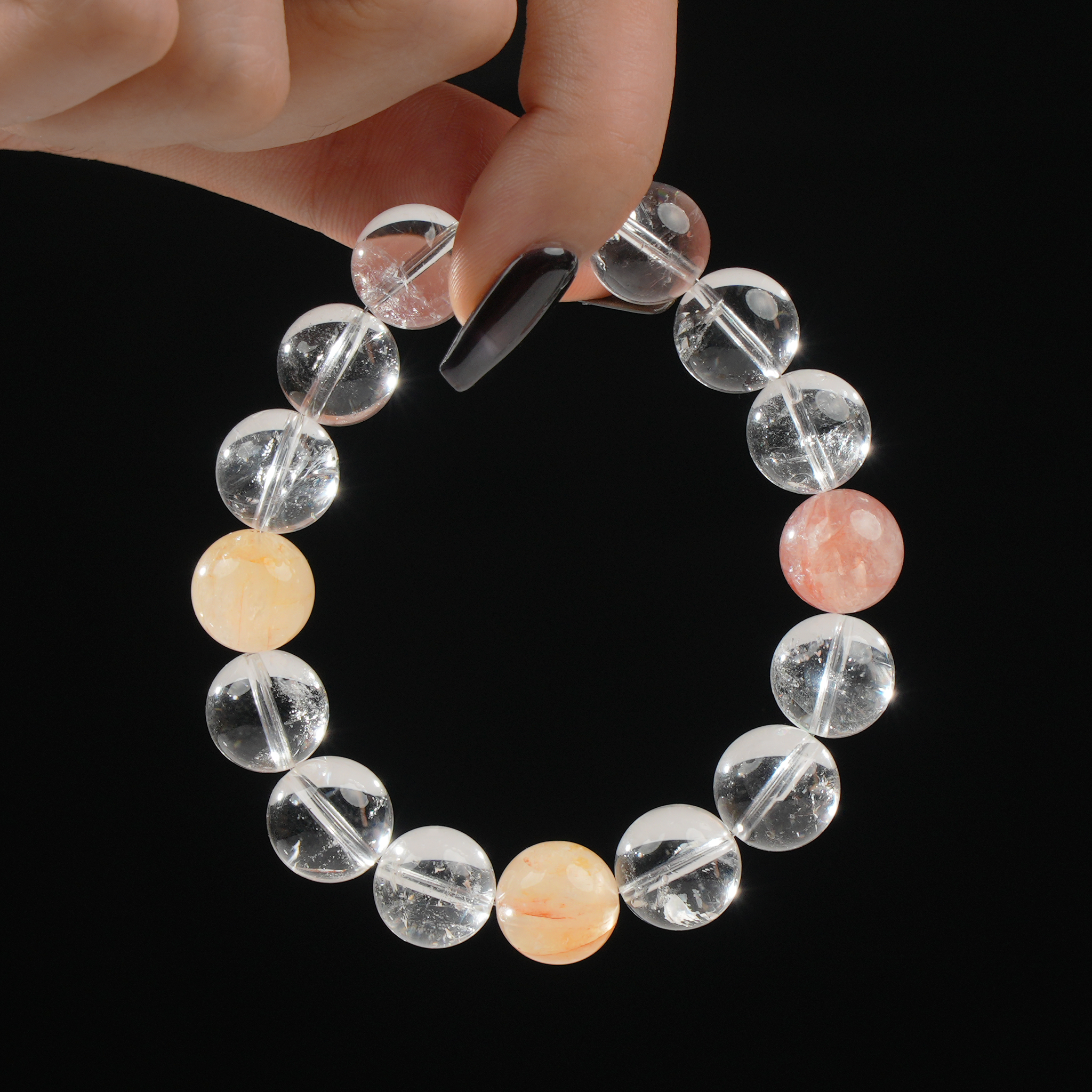 A close-up of a hand with long black nails holding a bracelet composed of Clear Quartz and Hematoid Quartz beads, set against a black background for contrast.