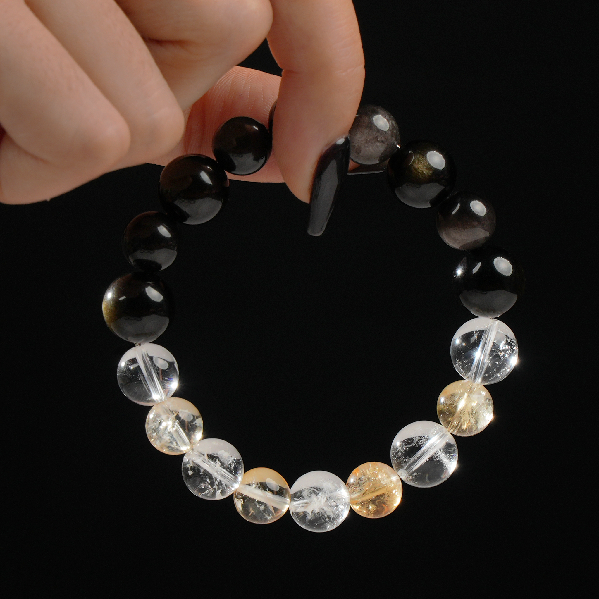 This photo captures a person gently holding a round gemstone bracelet between two fingers. The bracelet consists of polished 12mm beads in a gradient of tones: bright Clear Quartz, sunny Citrine, metallic Silver Obsidian, and dark glossy Obsidian. The stones’ textures and reflections are emphasized by the clean black backdrop and soft lighting.