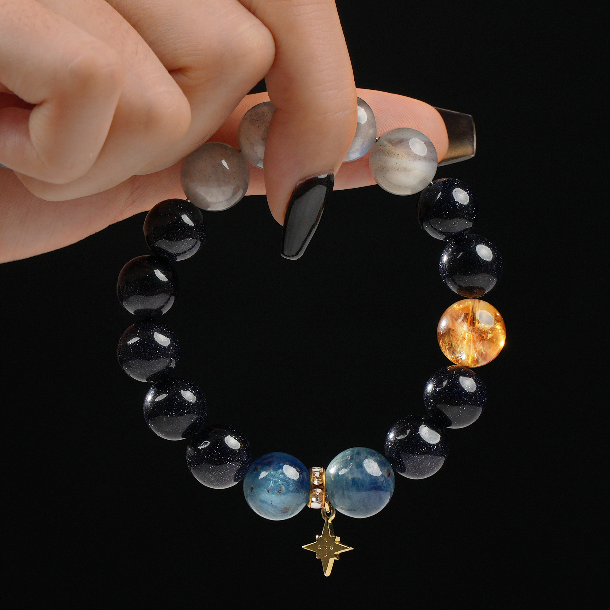 The bracelet displays a harmonious blend of gemstones, including soft blue Aquamarine, shimmering Moonstone, sparkling deep navy Blue Goldstone, and a radiant Citrine accent bead. A delicate gold star-shaped charm with a small crystal detail hangs between the Aquamarine beads. The bracelet is held between two fingers, highlighting its celestial and elegant design.