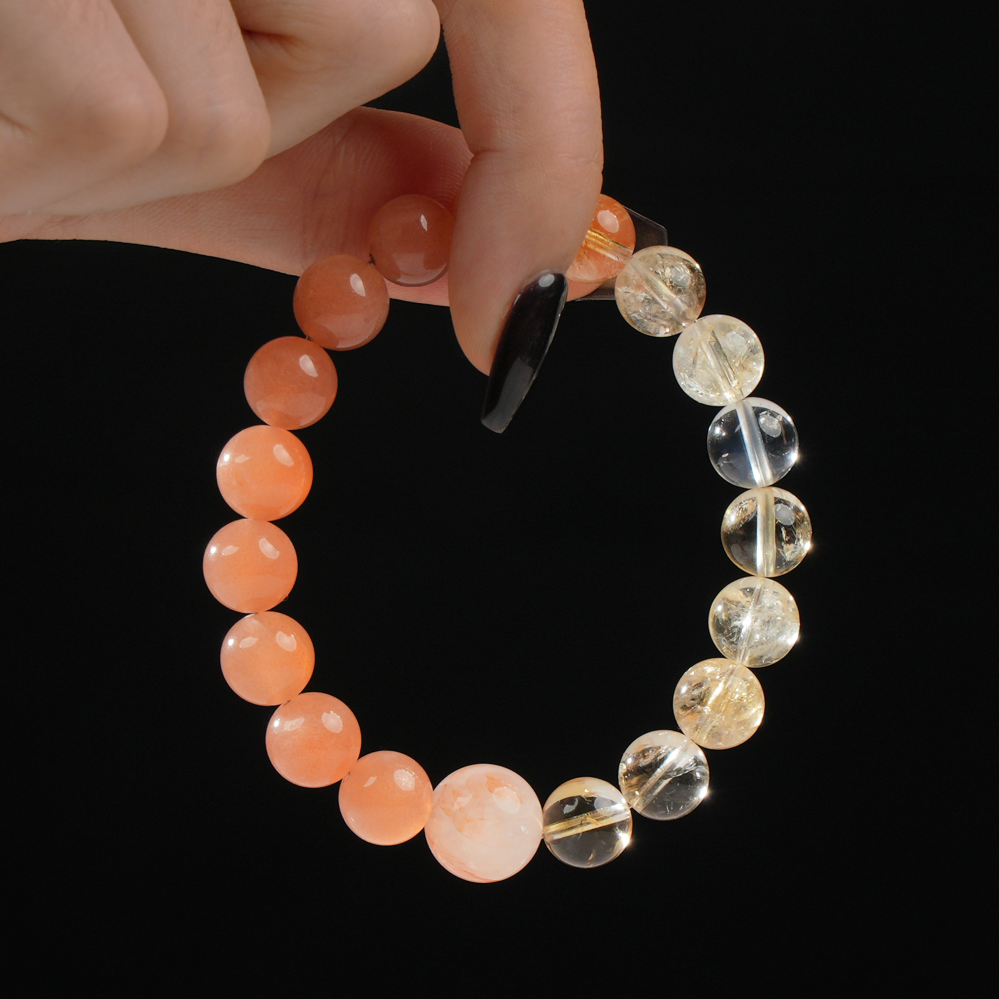 In this image, a hand delicately holds a round beaded bracelet made of natural gemstones. The bracelet features soft orange moonstone beads on one side and bright yellow citrine on the other, with a central bead of red rutilated quartz standing out for its internal coppery threads. The black background adds contrast to highlight the warm colors of the stones.
