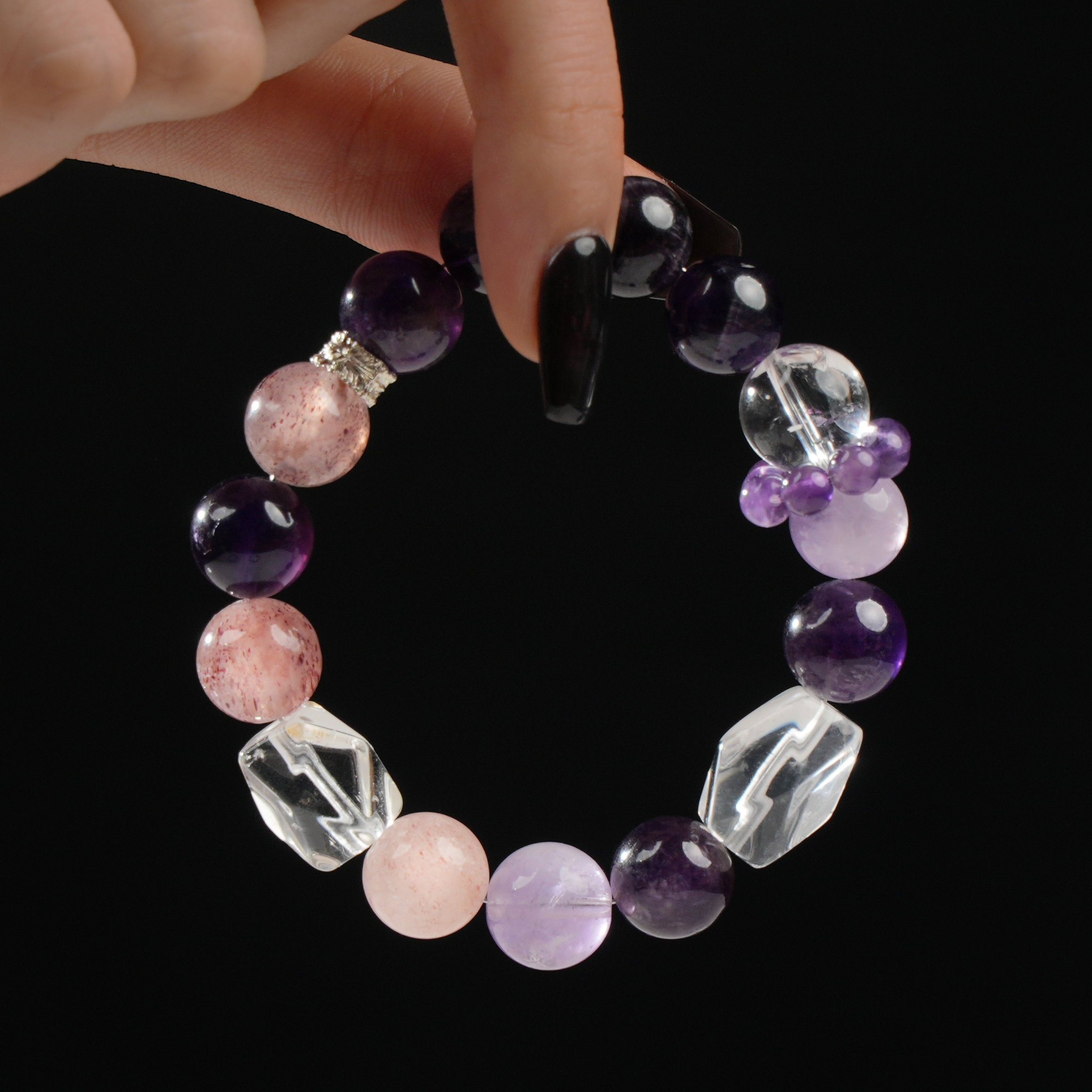 This bracelet showcases a rich combination of gemstones including deep purple Amethyst, soft pink Strawberry Quartz, and angular faceted Clear Quartz pieces. Accented with silver-tone spacers and a cluster of smaller round Amethyst beads, it balances texture and color beautifully. The bracelet is held between fingers, with its luminous stones contrasting against the dark background.