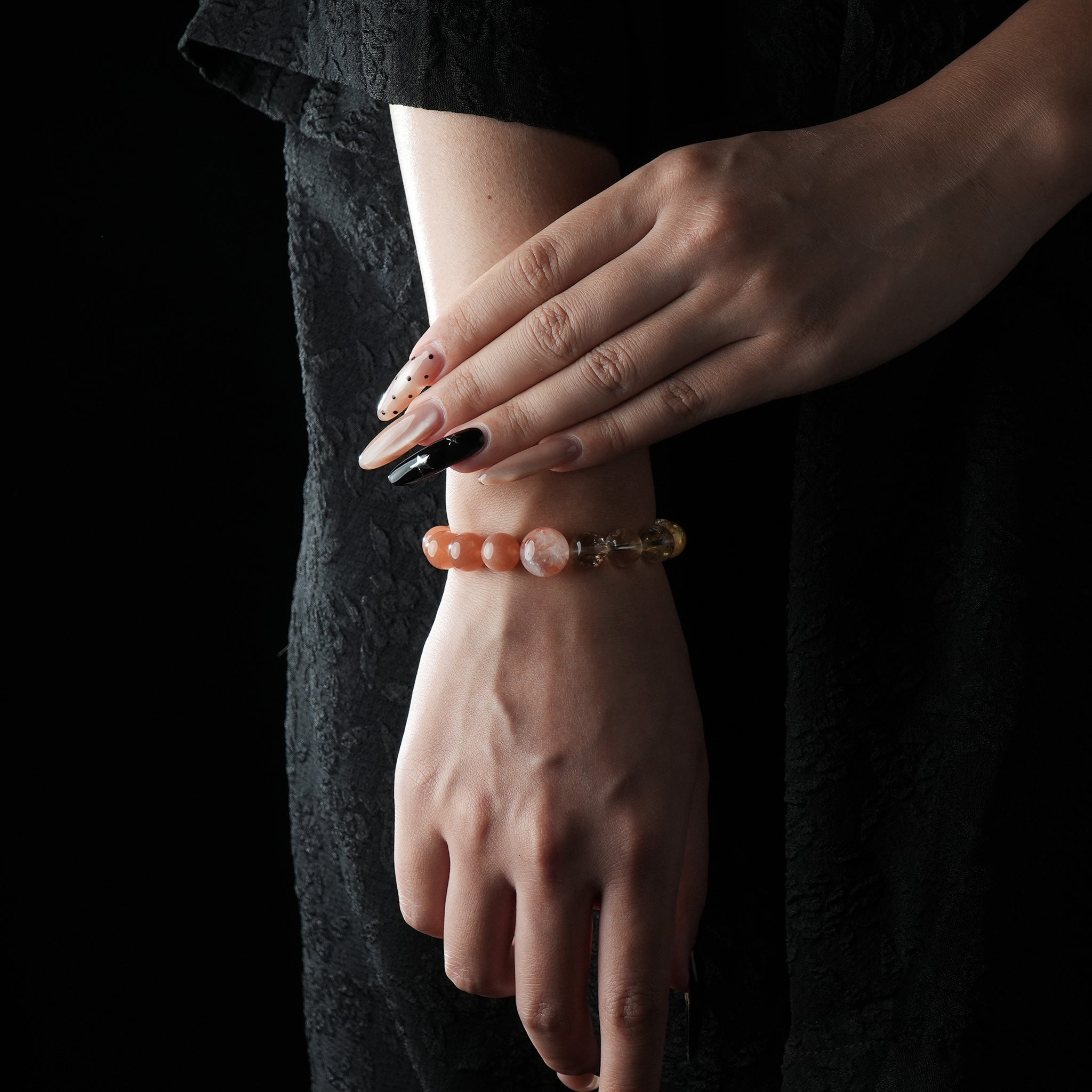 The image features a fashion model wearing the bracelet on their left wrist, with hands gently posed. The bracelet’s orange and golden tones complement the model’s dark, elegant outfit. The lighting subtly catches the gemstone reflections, giving the accessory a warm, radiant appearance.