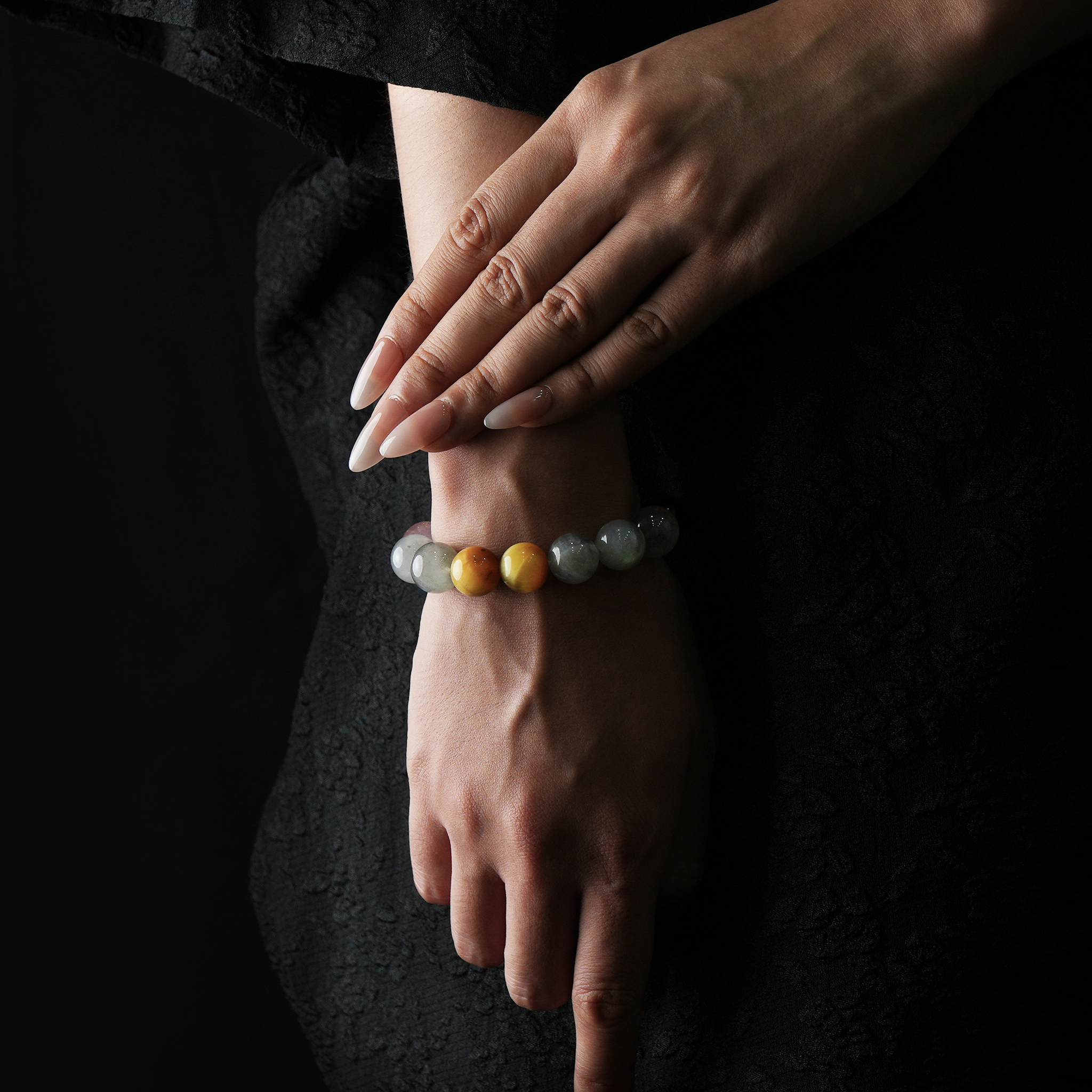 A close-up of a woman's hands elegantly resting on her wrist, showcasing a natural crystal bracelet composed of obsidian, strawberry quartz, grey moonstone, and yellow tiger’s eye beads. The woman wears a black textured dress, and the background is dark, highlighting the subtle glow and contrast of the stones.