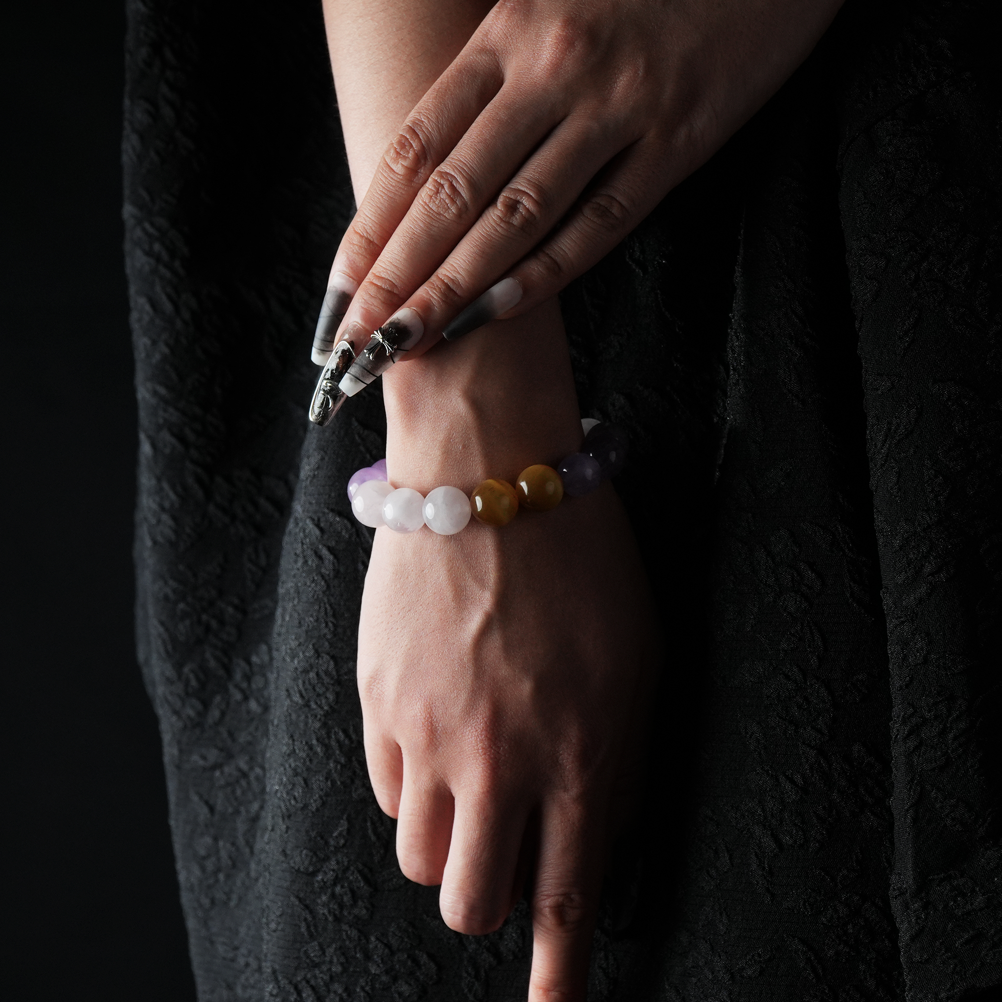A close-up of a hand wearing a beaded bracelet made of Amethyst, Rose Quartz, and Yellow Tiger Eye. The soft lavender and blush tones are accented by warm golden-brown beads, creating a gentle yet eye-catching color contrast against a black textured outfit.