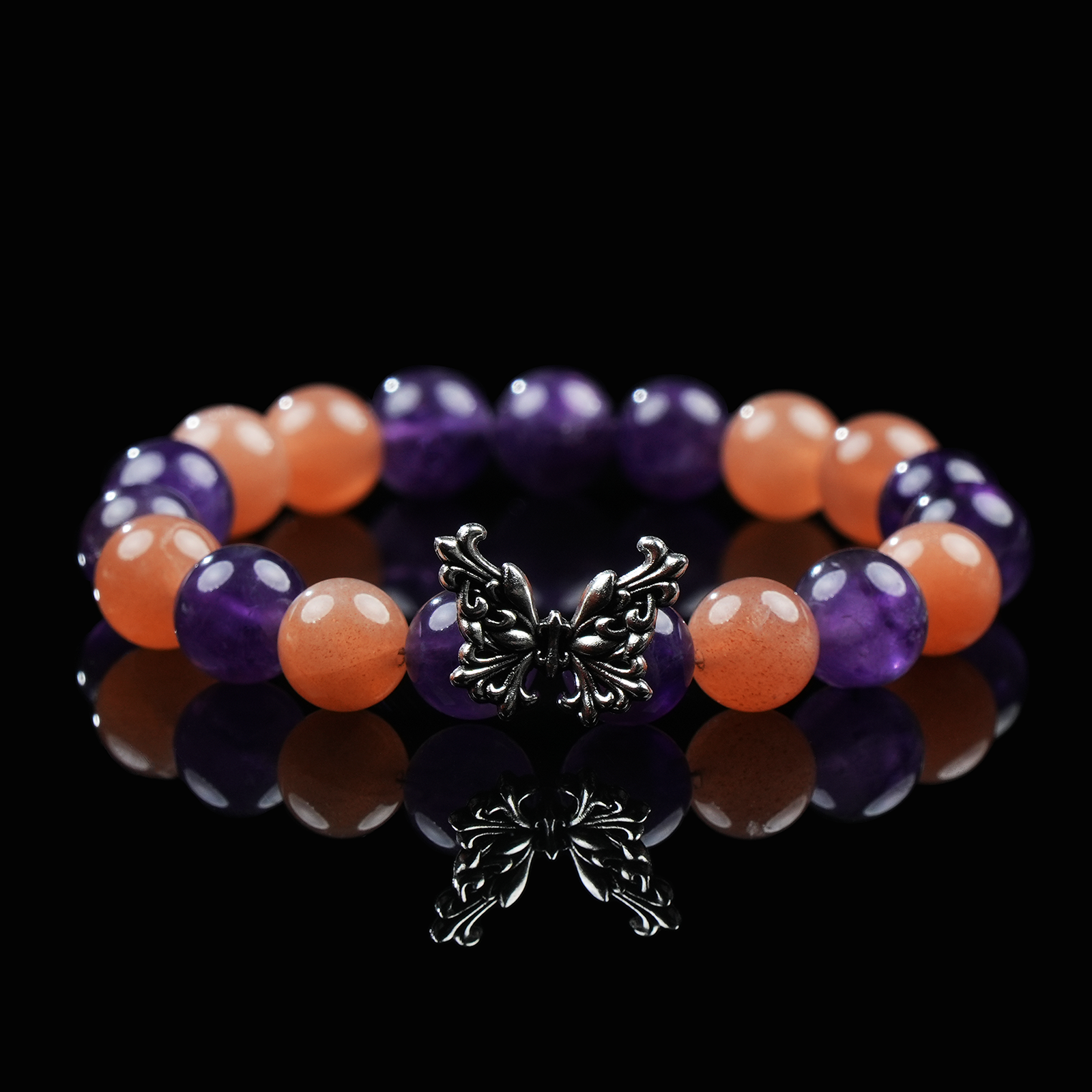 A vibrant beaded bracelet featuring alternating Amethyst and Orange Moonstone beads, centered with a detailed silver butterfly charm, placed on a glossy black reflective surface.