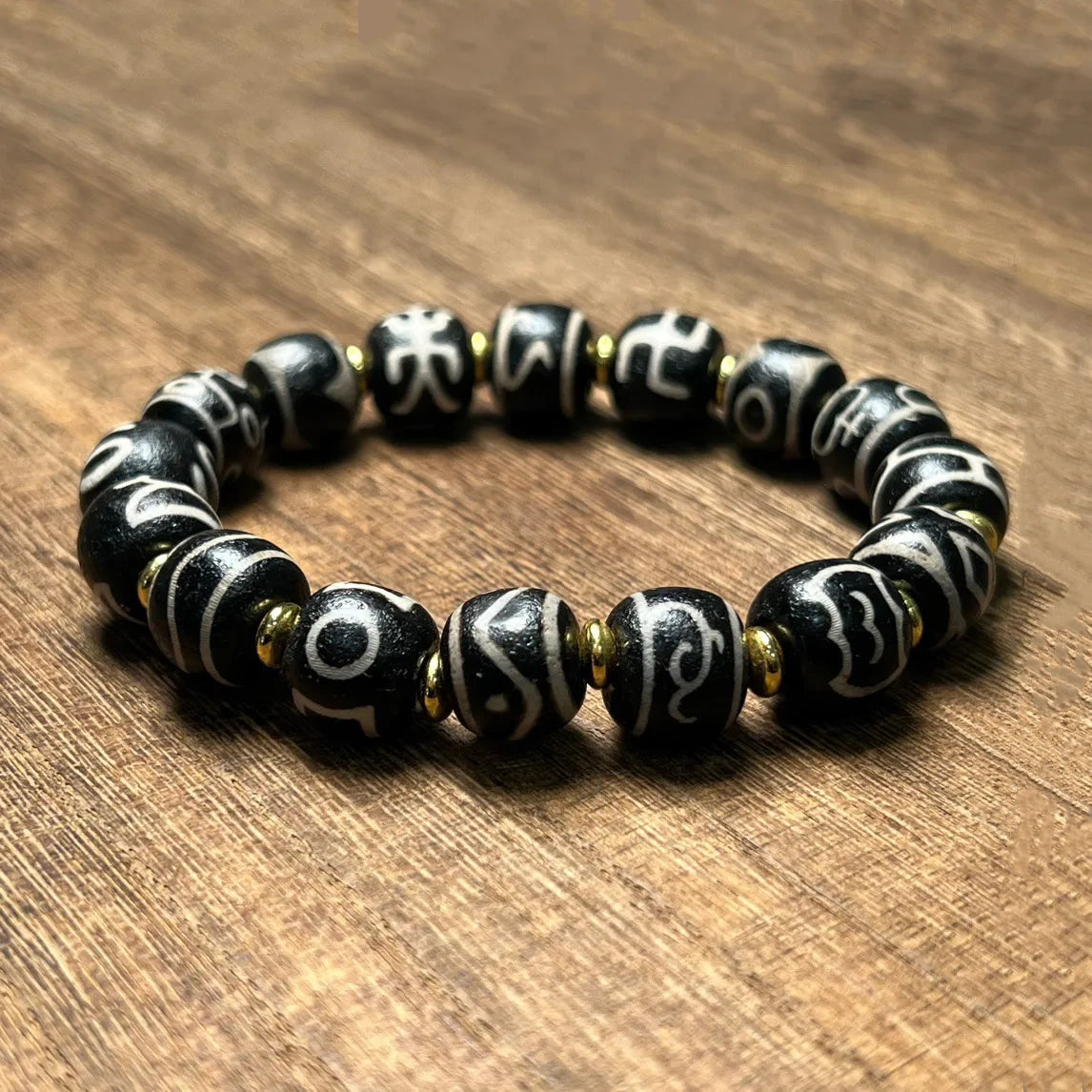 A Tibetan-style mantra bead bracelet displayed in a slightly angled view on a wood background. The bracelet features multiple black beads adorned with distinct white Tibetan symbols, separated by small gold spacers, representing a mix of spiritual symbolism and rustic elegance.