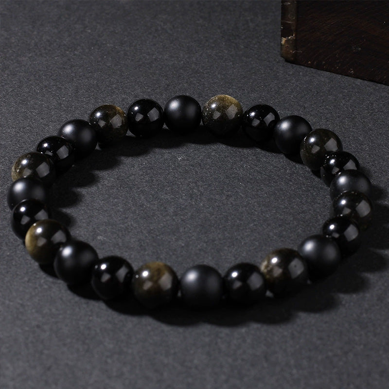 Bracelet laid flat on dark surface, composed of matte black agate and reflective gold sheen obsidian beads, offering strength and emotional shielding.