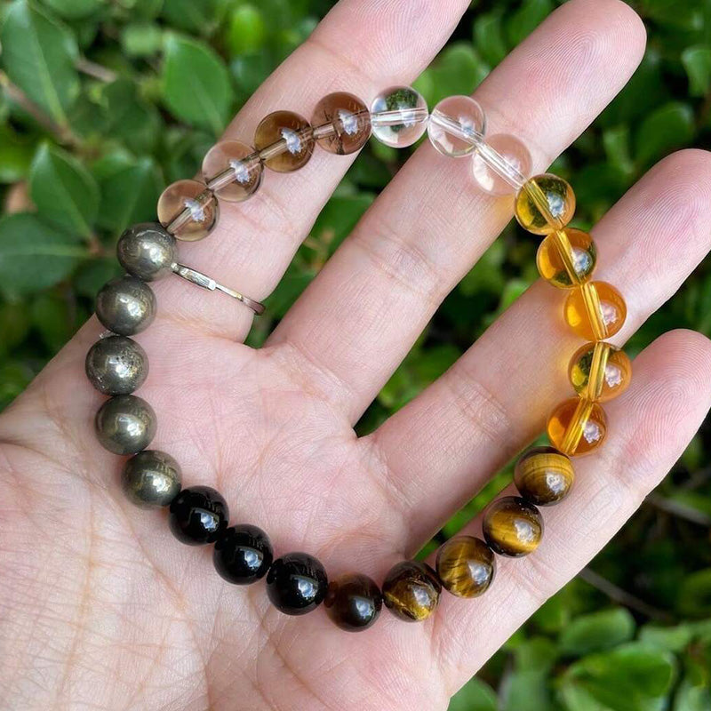 A natural light photo showing the bracelet resting on a hand, revealing the shine and texture of clear quartz, tiger’s eye, and black stones under outdoor lighting.