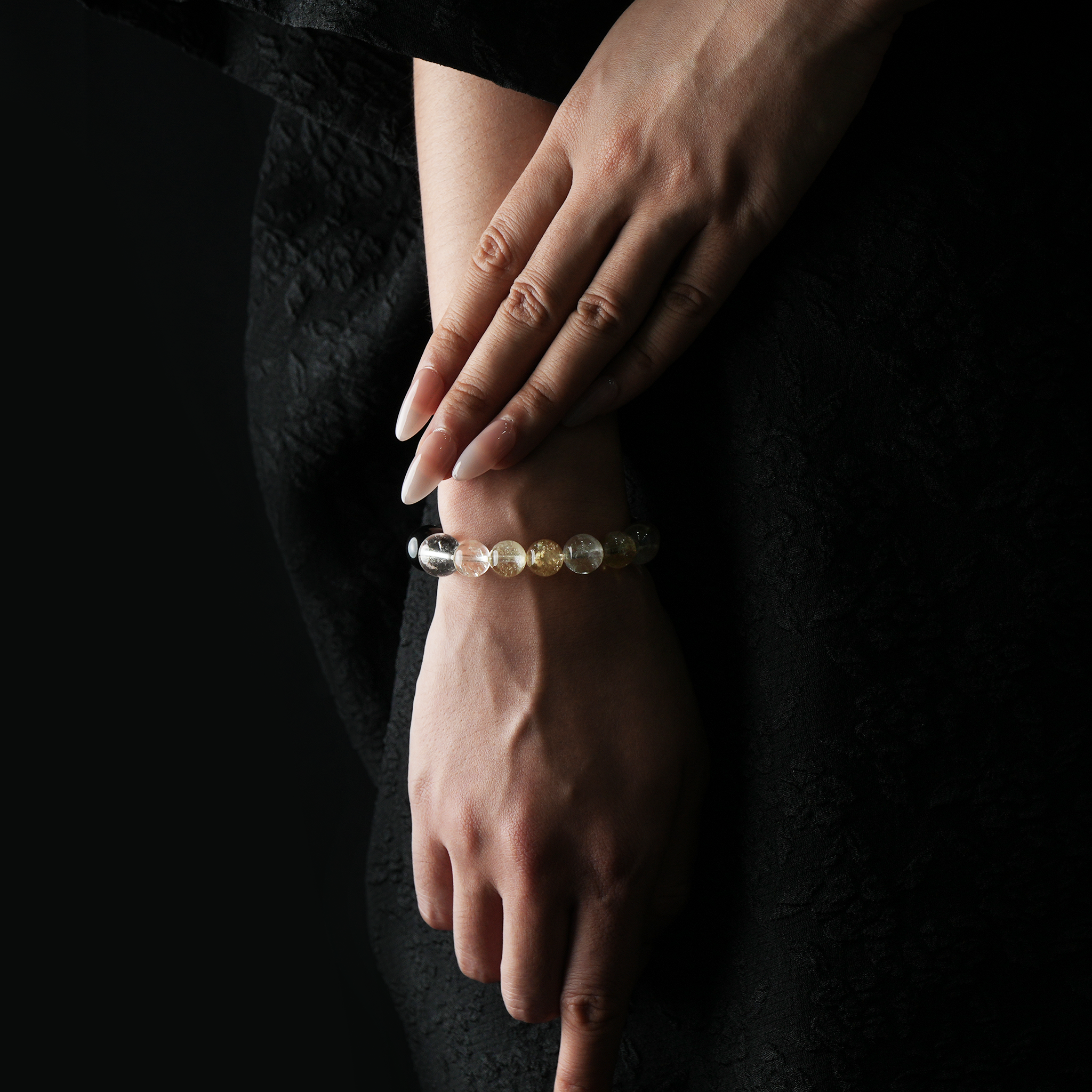 A woman’s wrist adorned with a luminous crystal bracelet featuring clear quartz, golden citrine, and silver-toned obsidian beads, shown against a black textured dress.
