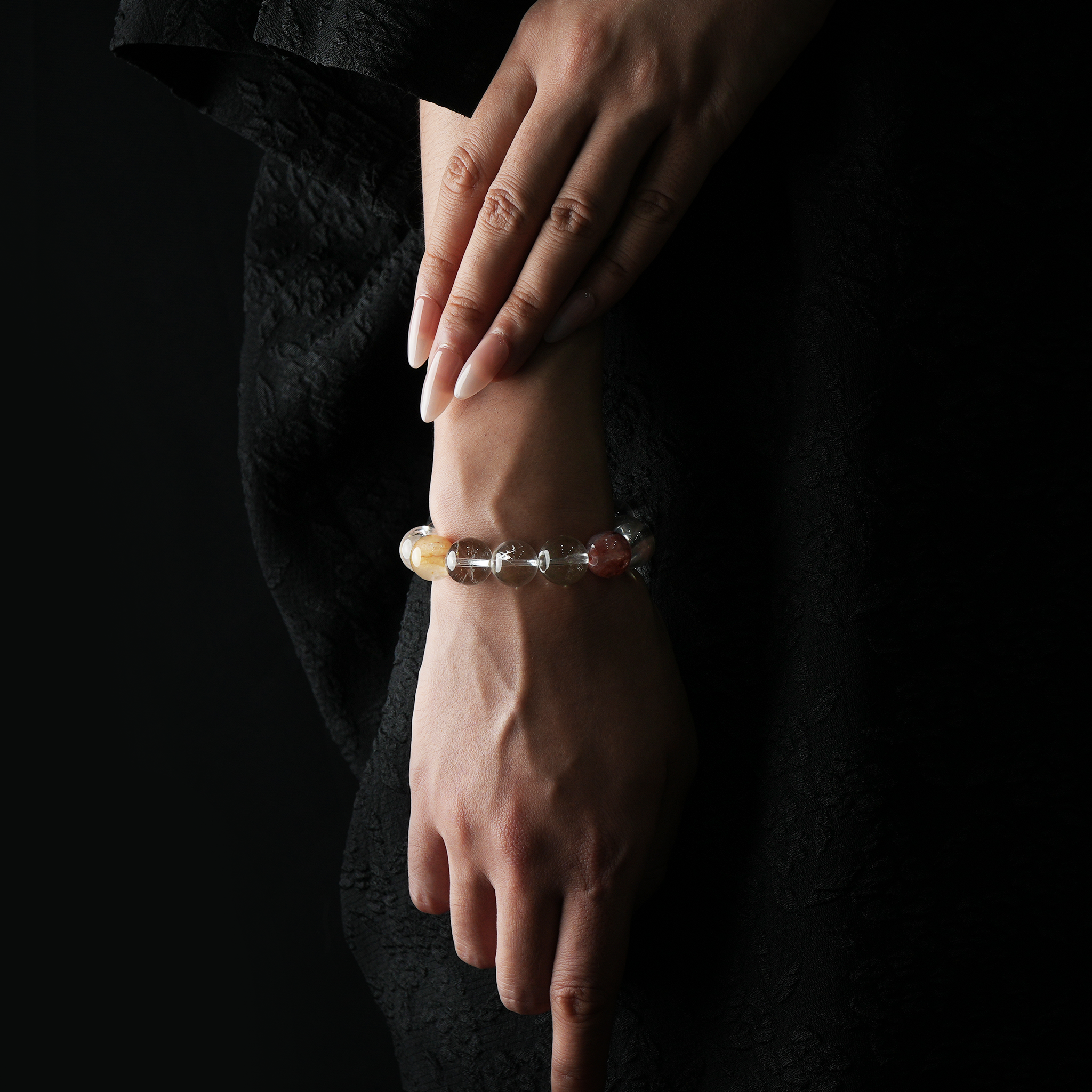 A woman's hand wearing a translucent crystal bracelet featuring hematoid quartz and clear quartz, photographed against a black outfit with soft lighting.