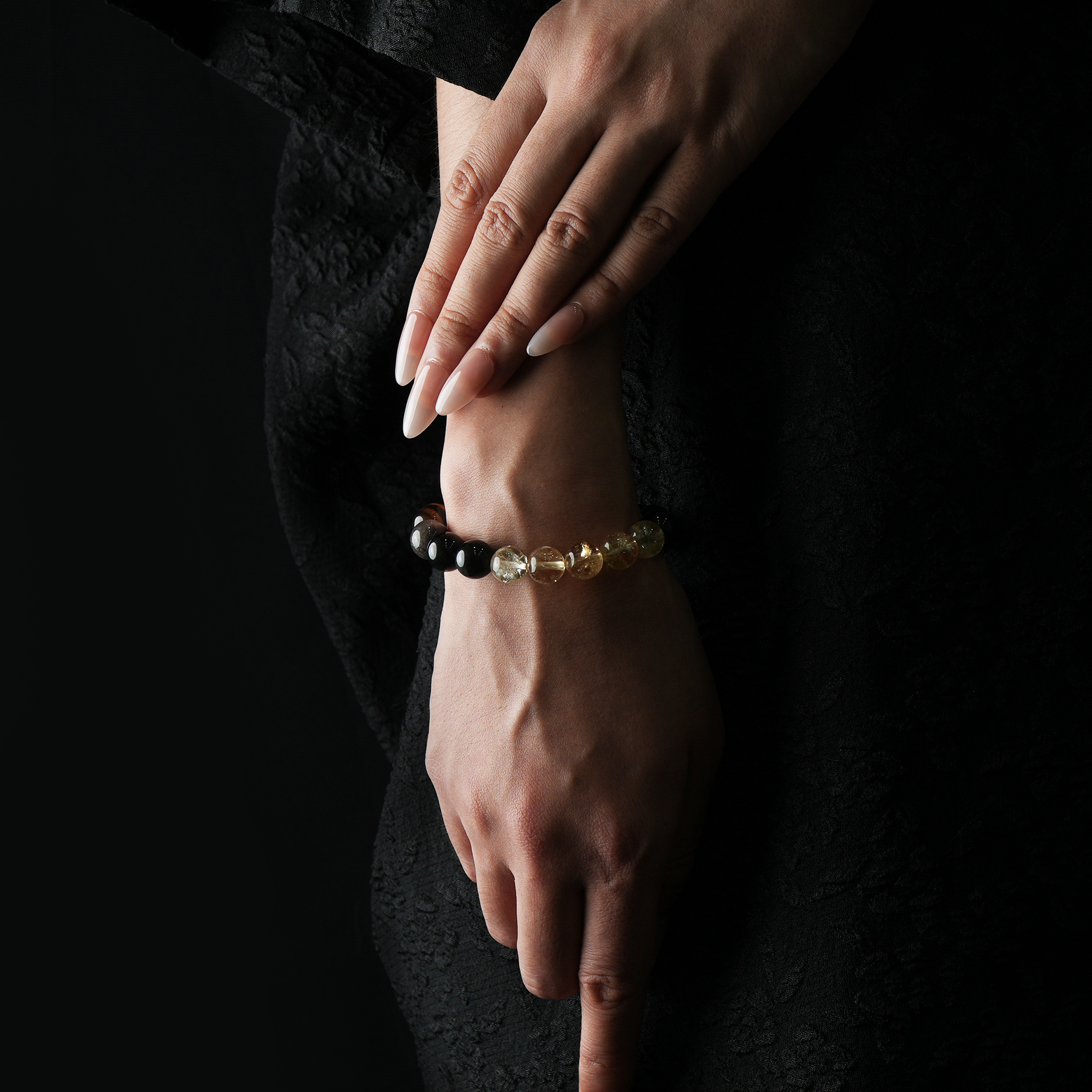 A woman’s hand wearing a golden-toned crystal bracelet featuring tiger’s eye, citrine, and silver sheen obsidian, set against a black outfit.
