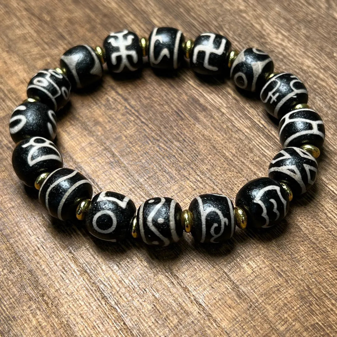 A circular arrangement of a handcrafted Tibetan Dzi bead bracelet resting on a wooden tabletop. The black beads, each inscribed with unique white script or symbols, are interspaced with golden spacers. The image emphasizes the symmetry, spiritual meaning, and earthy tone of the design.