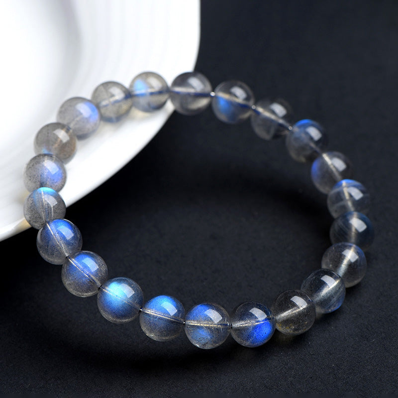 Gray moonstone bracelet casually resting near a white dish, with multiple beads showing distinct flashes of blue under soft lighting.