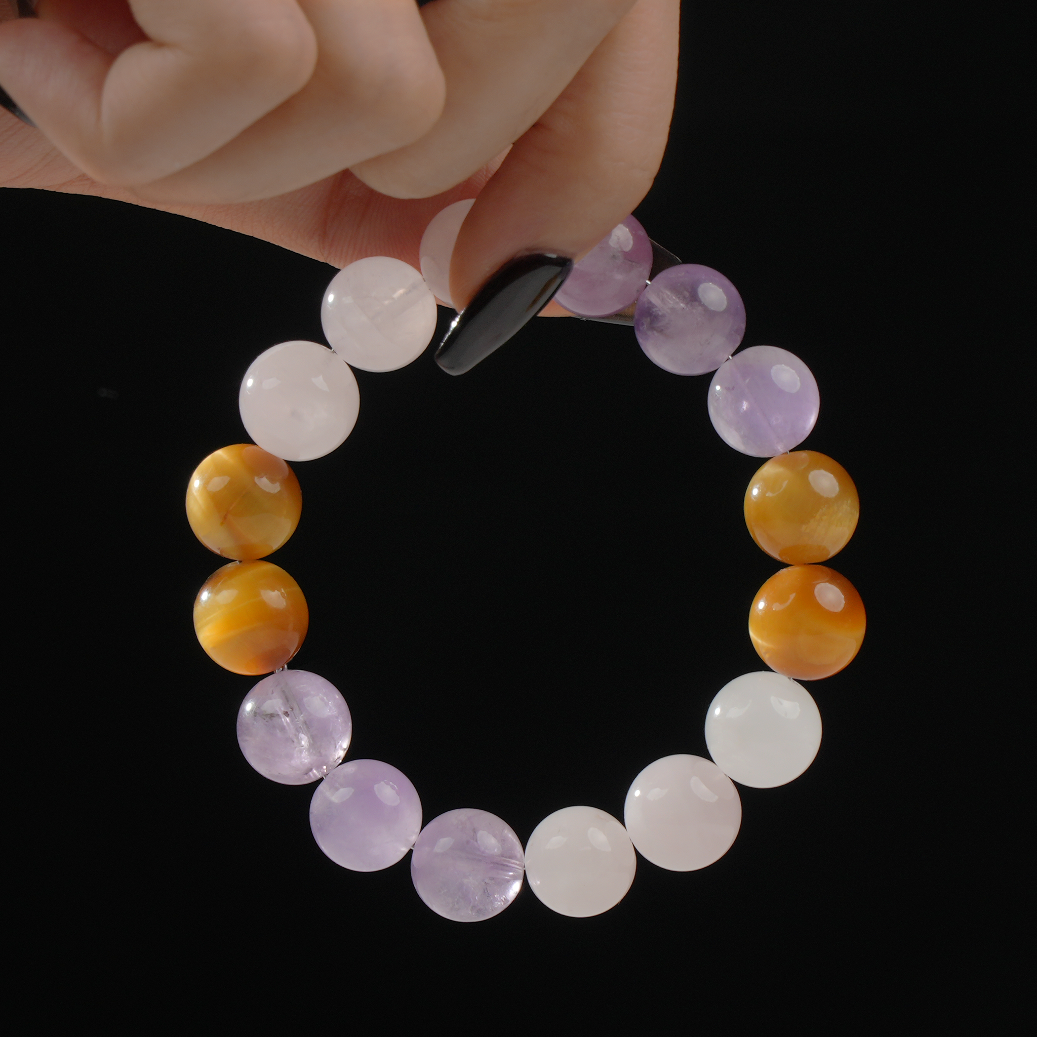 A close-up image of a hand gently holding a circular crystal bracelet made of polished white quartz, amethyst, and golden-yellow tiger’s eye beads, displayed against a black background. The colors form a soft, elegant gradient that highlights the natural luster of each stone.