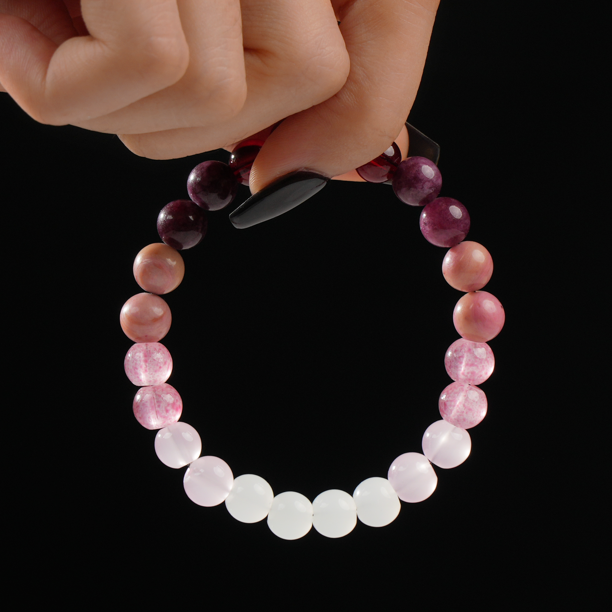 A hand displays a gemstone bracelet featuring a smooth gradient of natural Rose Quartz, Strawberry Quartz, and Garnet beads. The colors shift elegantly from soft white to pink and deep wine red, set against a black background that enhances the stones' natural shine and clarity.