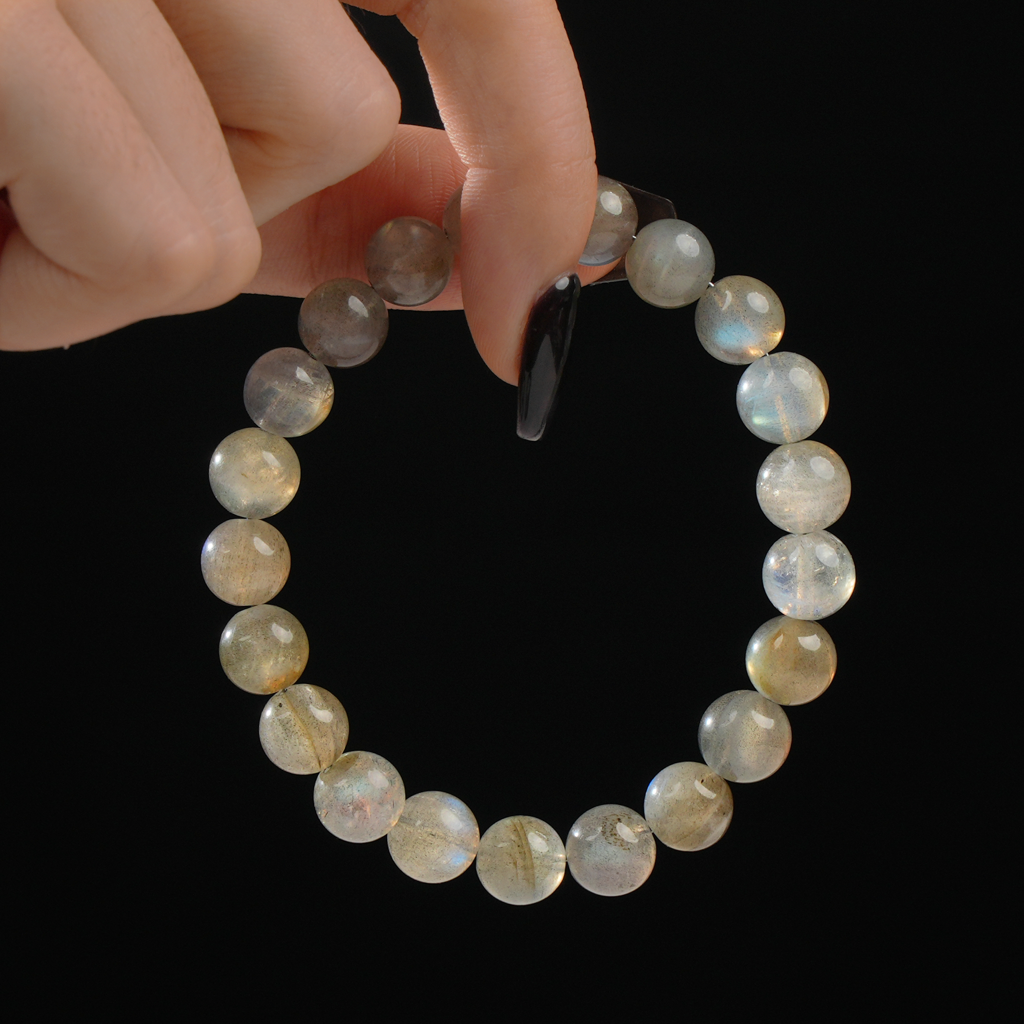 A close-up image of a hand holding a round bracelet composed of shimmering 12mm grey moonstone beads. The beads exhibit a soft glow and natural variations in tone, with hints of blue and gold flash under light. The bracelet is shown against a solid black backdrop, highlighting the mystical sheen of the moonstones.