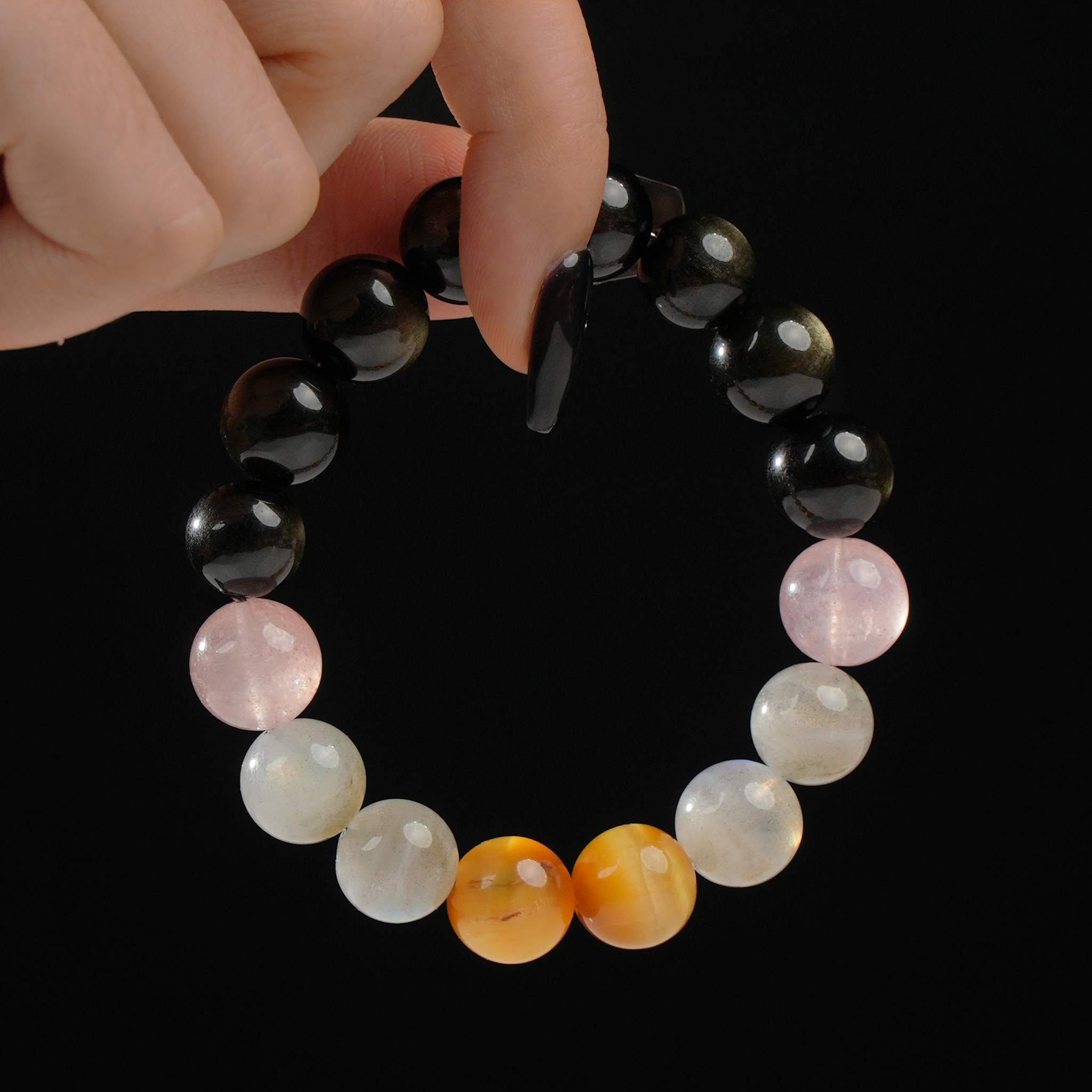 A close-up of a hand holding a round beaded bracelet, featuring glossy black Obsidian stones, soft pink Strawberry Quartz, shimmering Grey Moonstone, and two warm golden Tiger’s Eye beads. The stones are smooth and polished, arranged in a symmetrical gradient to create balance and elegance.