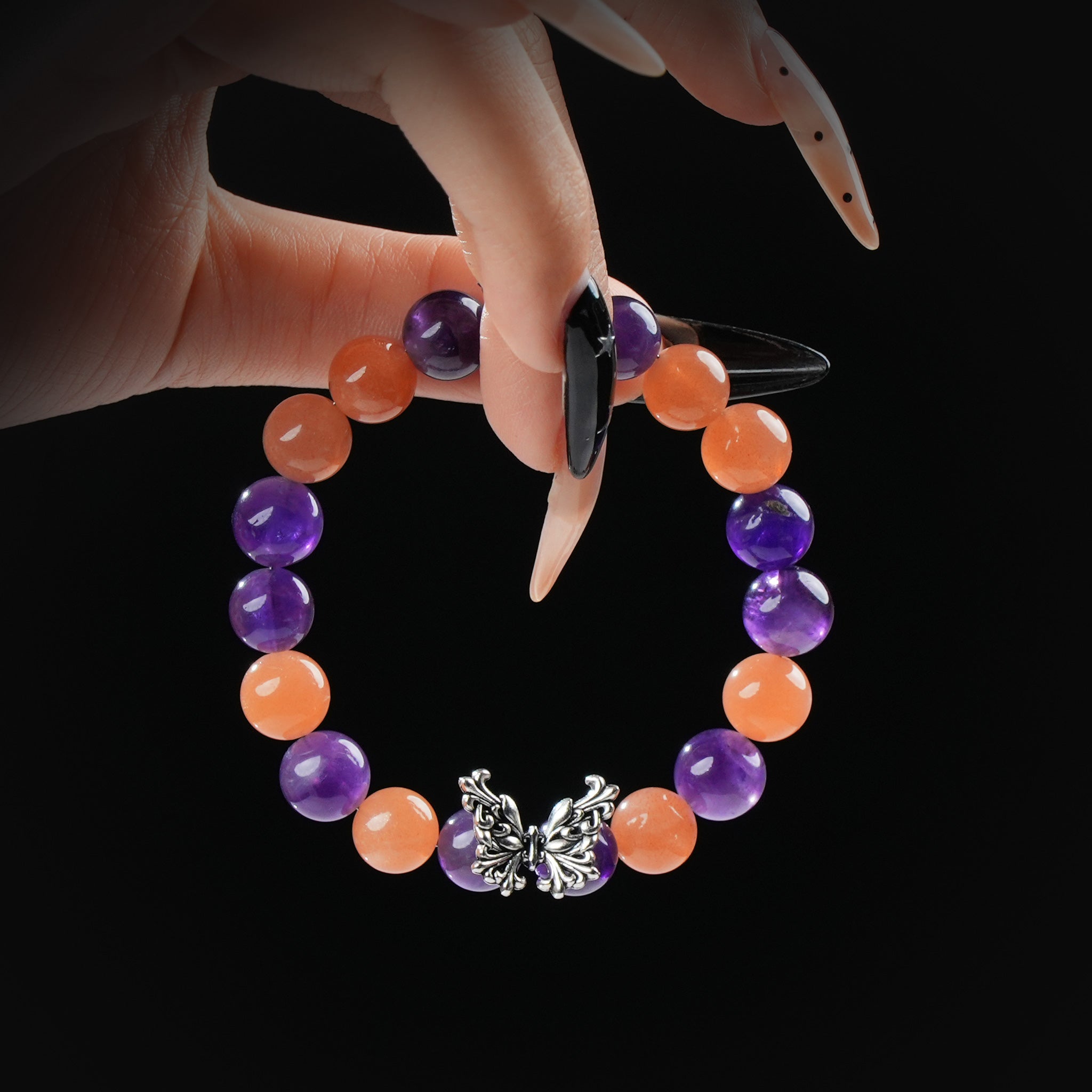 A close-up of a hand with long, manicured nails holding a beaded bracelet made of alternating Amethyst and Orange Moonstone stones, featuring a detailed silver butterfly charm at the center.