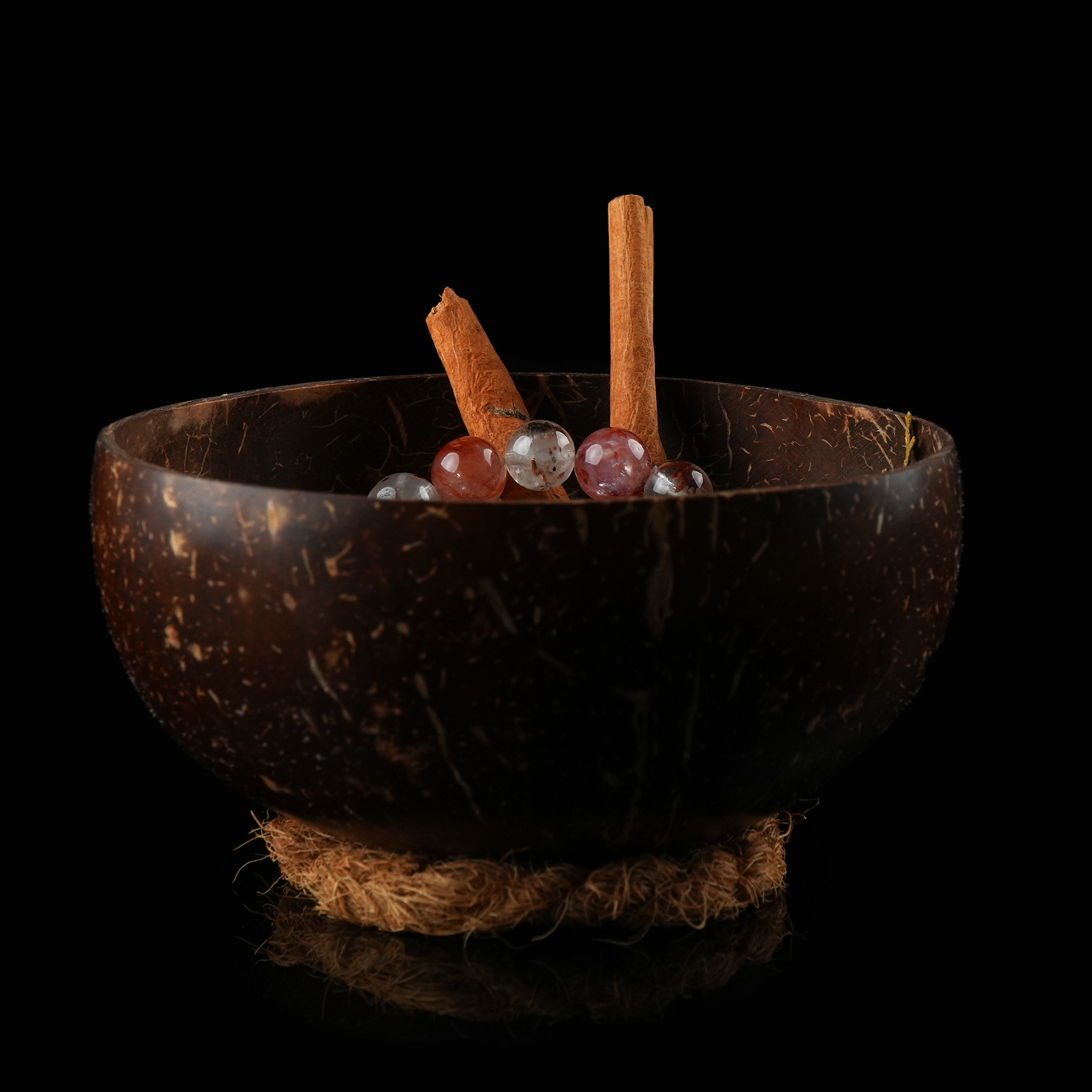 A small coconut bowl on a rope base, containing herbs and a gemstone bracelet under soft lighting.