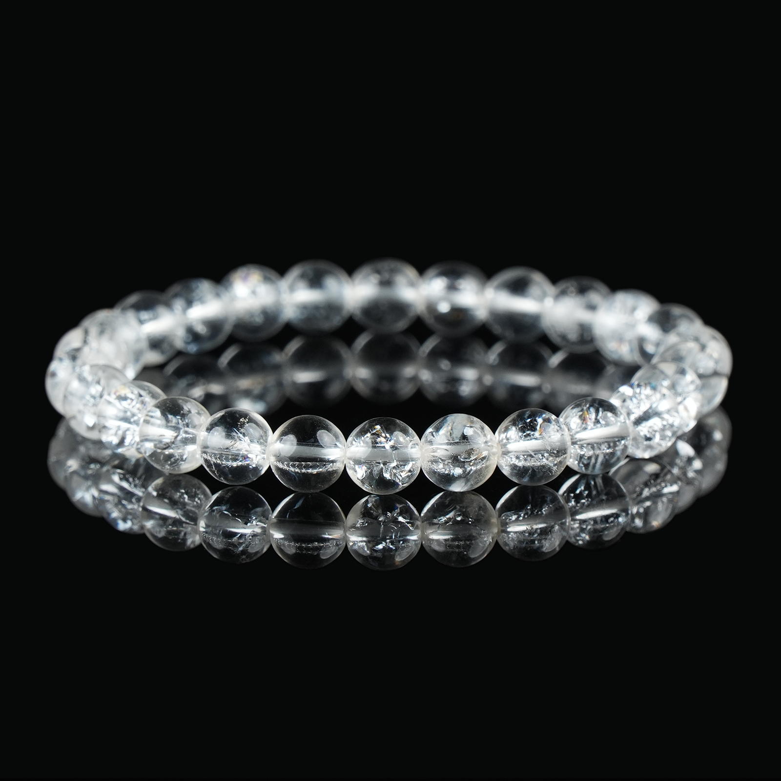 A clear quartz bracelet on a black reflective surface, highlighting the transparency and inner fractures of the crystal beads.