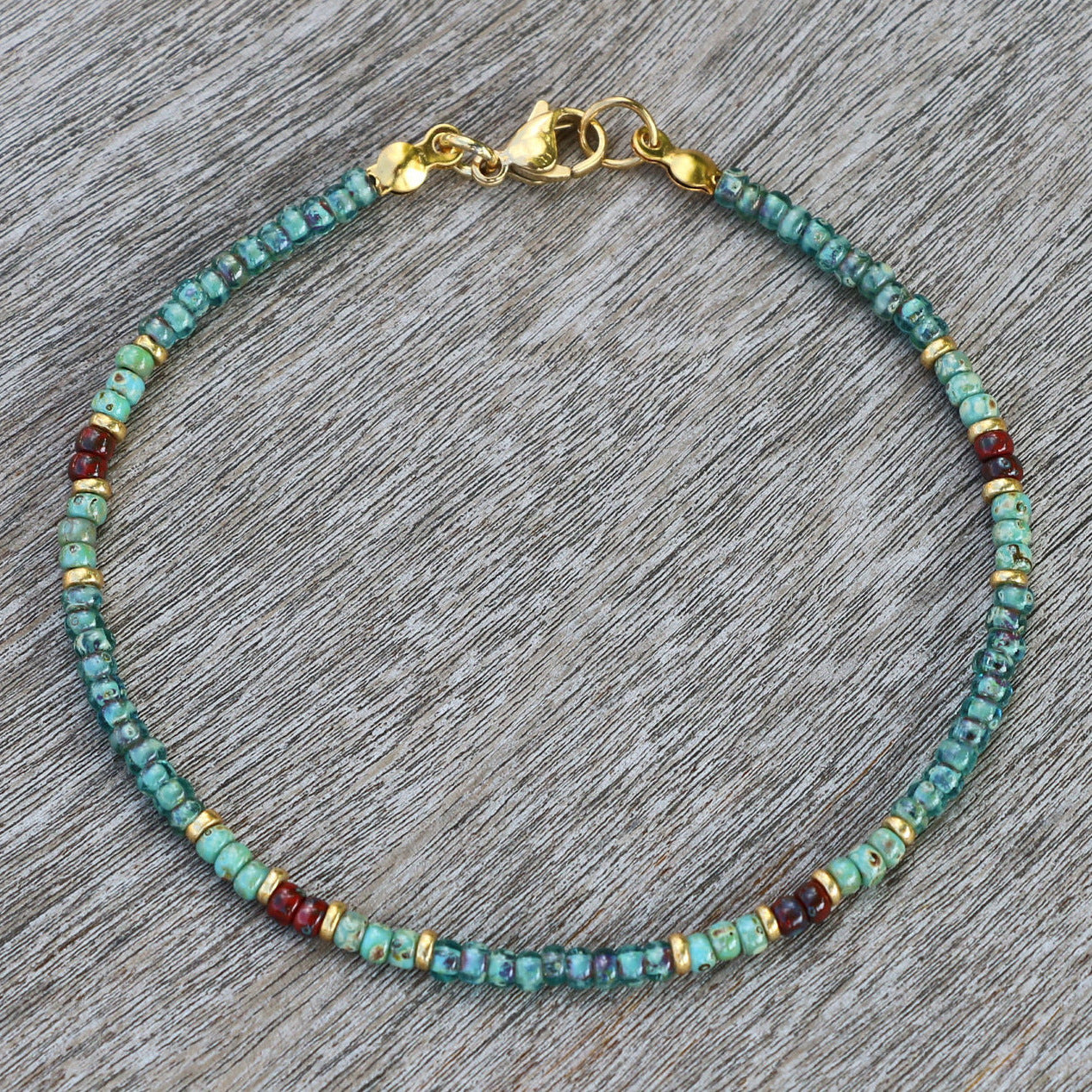 A close-up of a thin turquoise anklet with red, gold, and blue accents on a textured wooden background.
