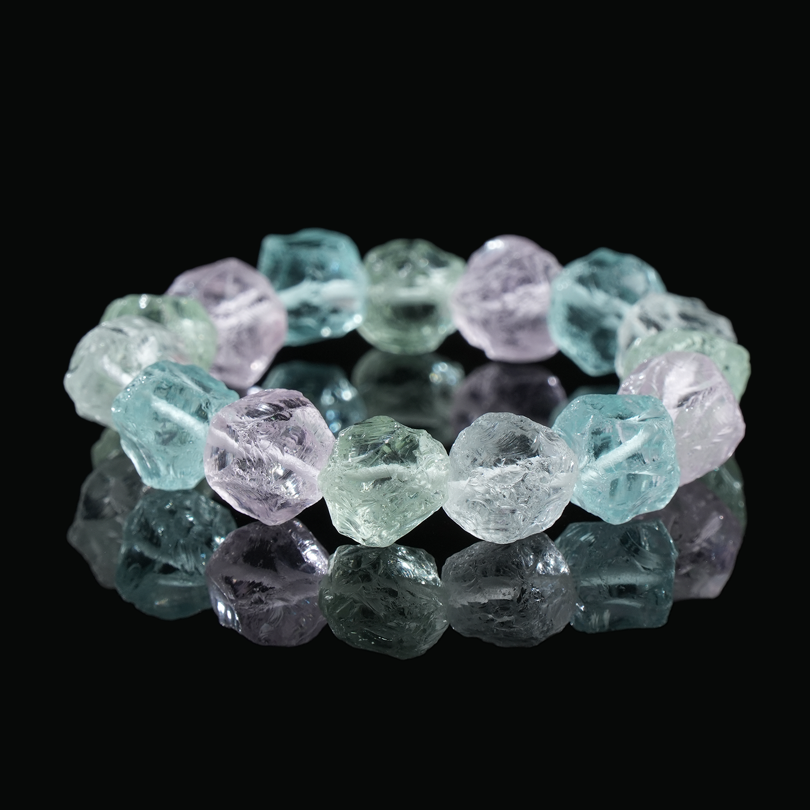 Close-up of a natural raw crystal bracelet, featuring irregular crystal shapes with shades of green, pink, and clear hues, reflecting light on a dark surface.