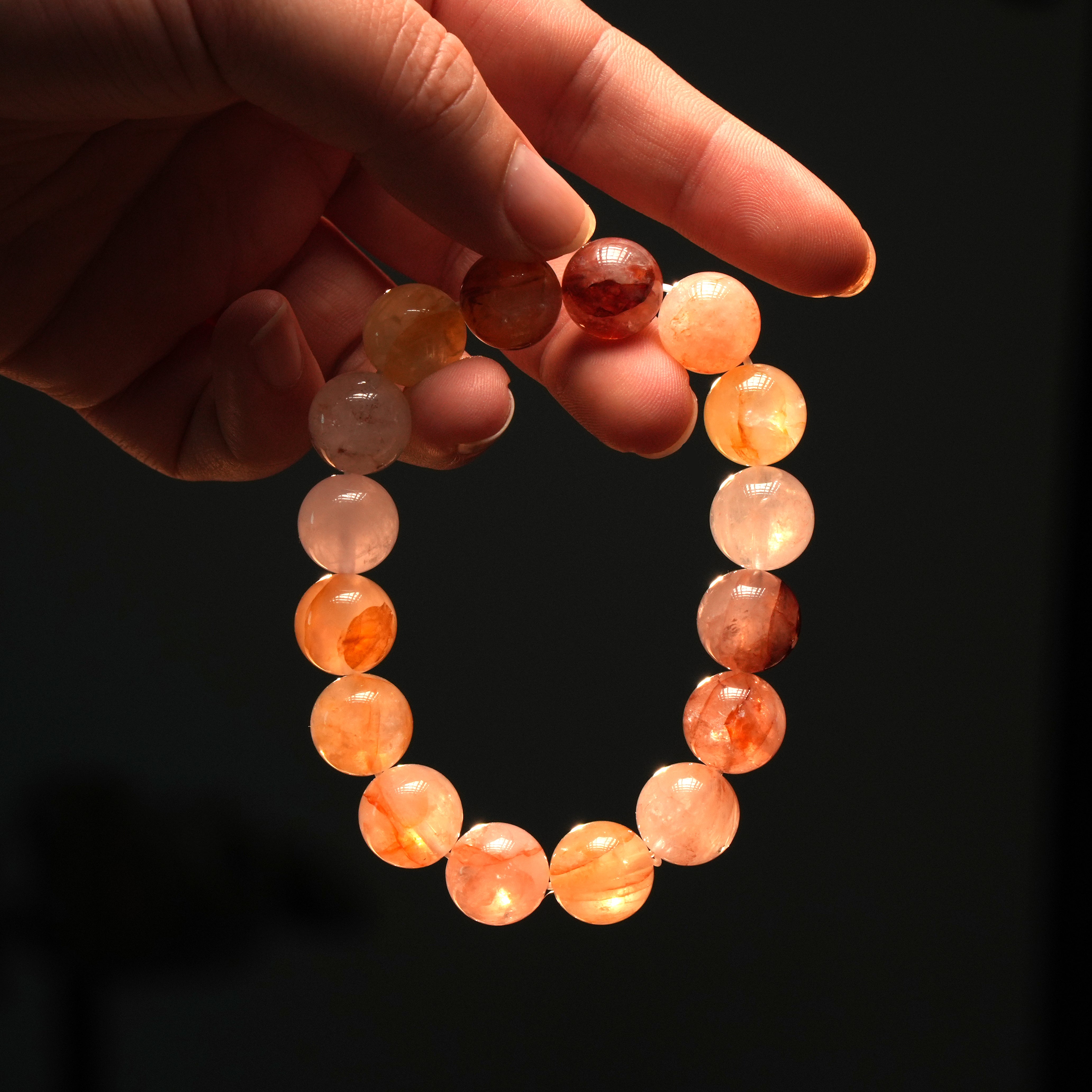 Side-lit image of a hand holding red rutilated quartz bracelet, highlighting its rich spectrum of fiery tones and the energizing presence it radiates.