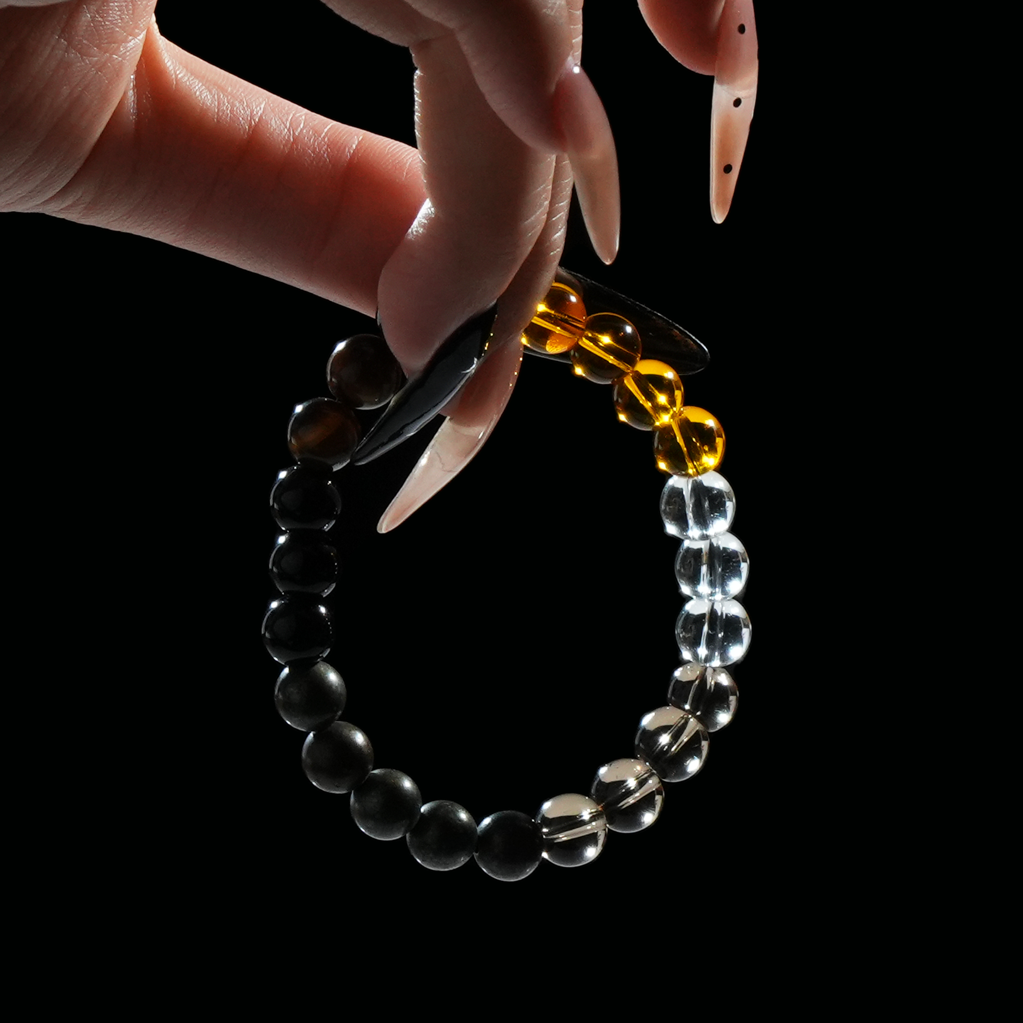 A hand gently holding a beaded bracelet featuring obsidian, clear quartz, and golden tiger’s eye stones, creating a striking contrast against the dark background.