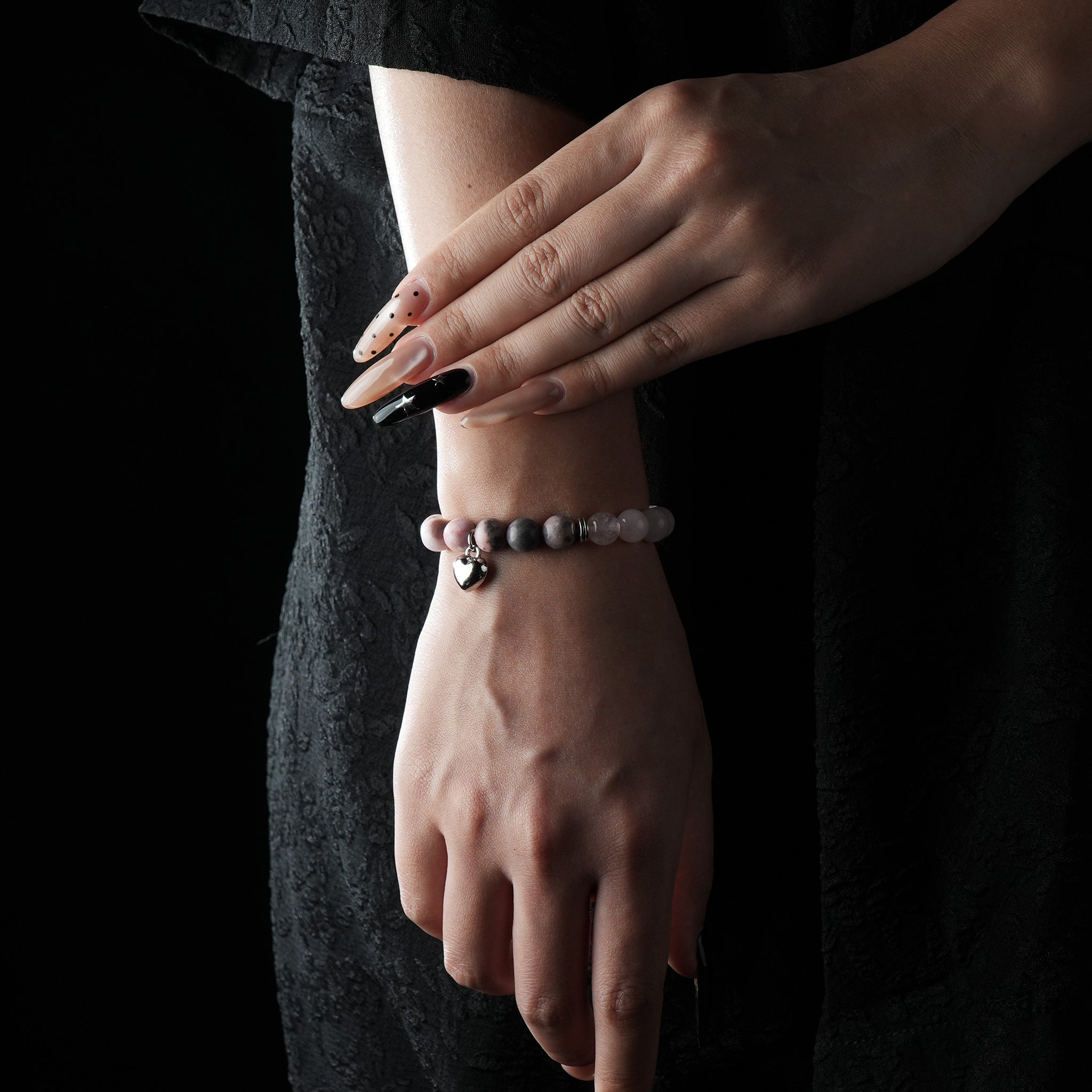 A delicate rose quartz beaded bracelet with a silver heart charm, worn on the wrist, styled with a black outfit.