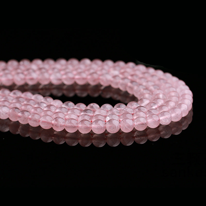 This image shows multiple strands of rose quartz beads arranged in a loose circular loop. The soft reflection on the black surface enhances the delicate pink color and smooth finish of each round bead.