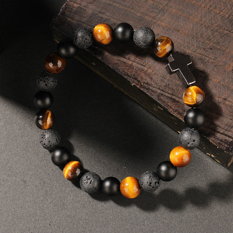 Bracelet resting on textured wood, composed of tiger’s eye, black obsidian, and porous lava beads, anchored by a silver cross charm.