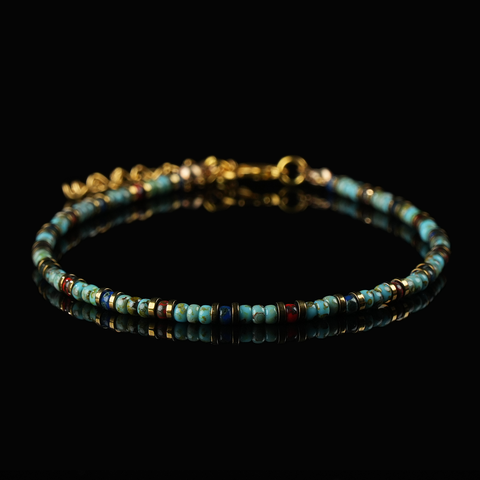 A close-up of a turquoise crystal anklet with colorful beads and gold clasp, displayed on a black background.