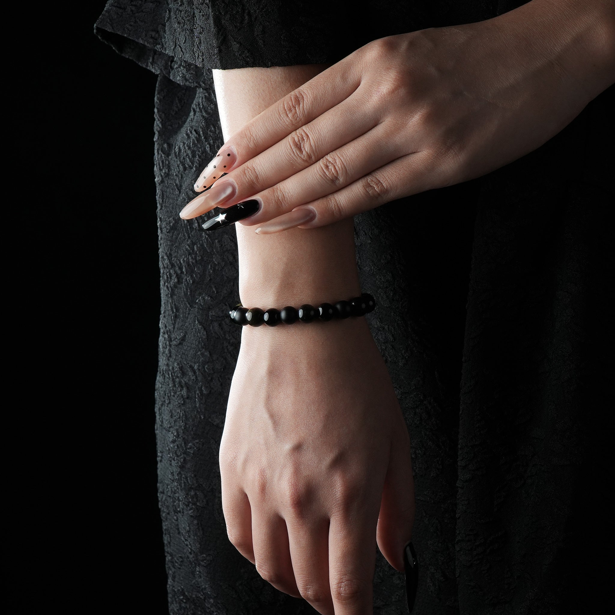 A close-up of a model’s wrist adorned with a simple black obsidian beaded bracelet, styled with a black lace dress and long manicured nails.