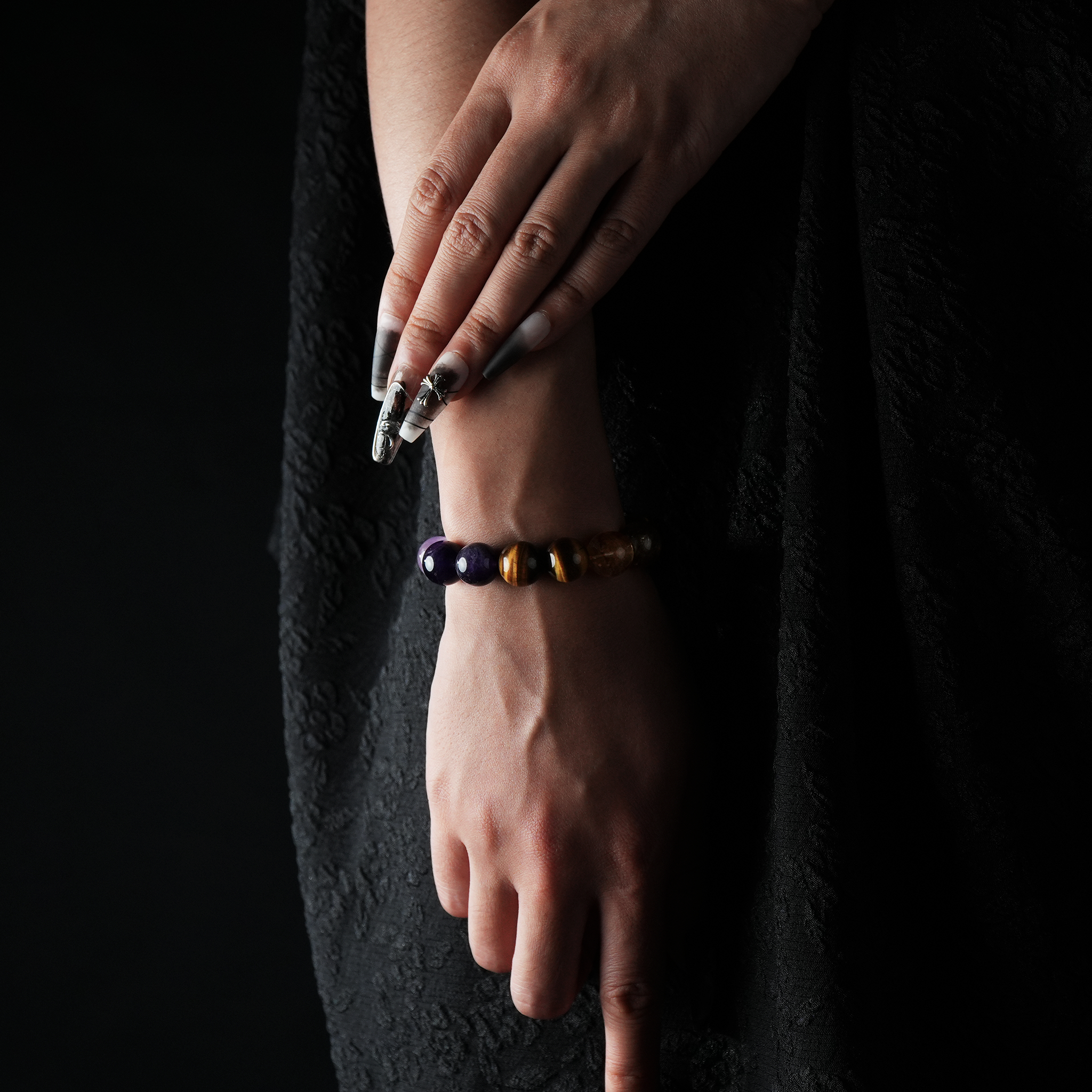 A stylish model with manicured nails wearing a gemstone bracelet featuring purple amethyst and golden tiger eye beads, contrasted against a black textured outfit and dark backdrop