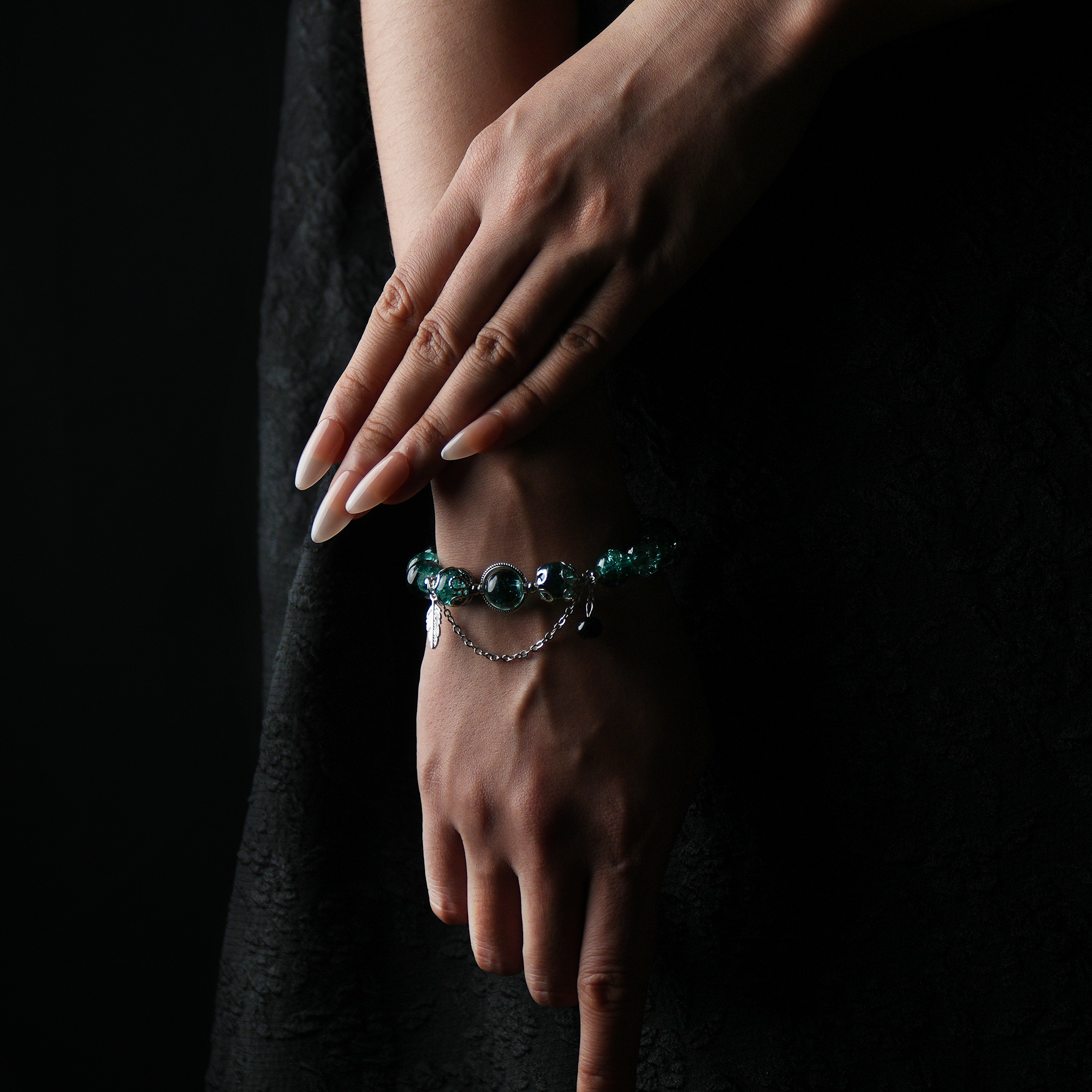 A wrist adorned with a dark green cracked energy bead bracelet featuring a silver feather charm and dainty chain, embodying forest strength and intuitive calm.