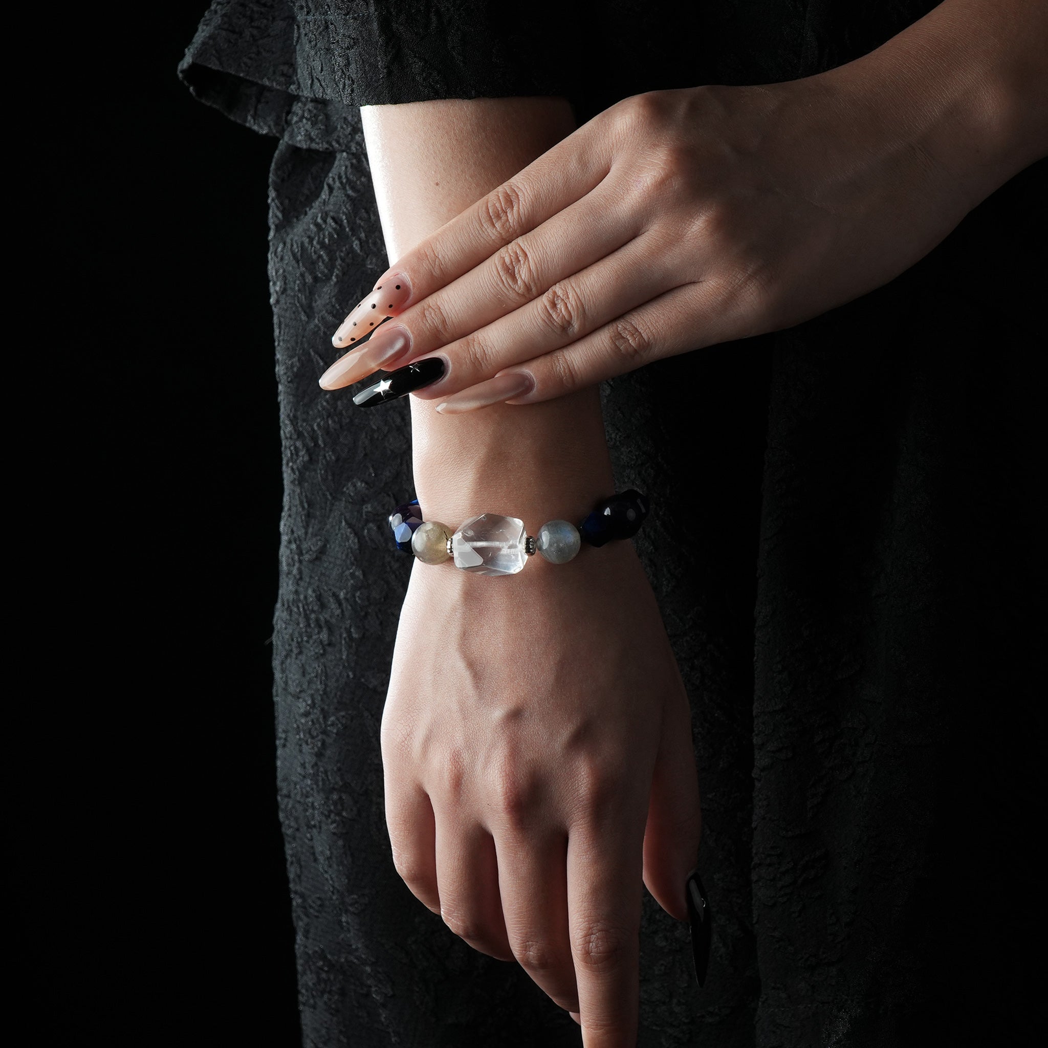 A close-up of a hand modeling a beaded bracelet featuring deep blue lapis lazuli, purple amethyst, glowing moonstone, and a large faceted clear quartz centerpiece, paired with a black textured outfit and long manicured nails.