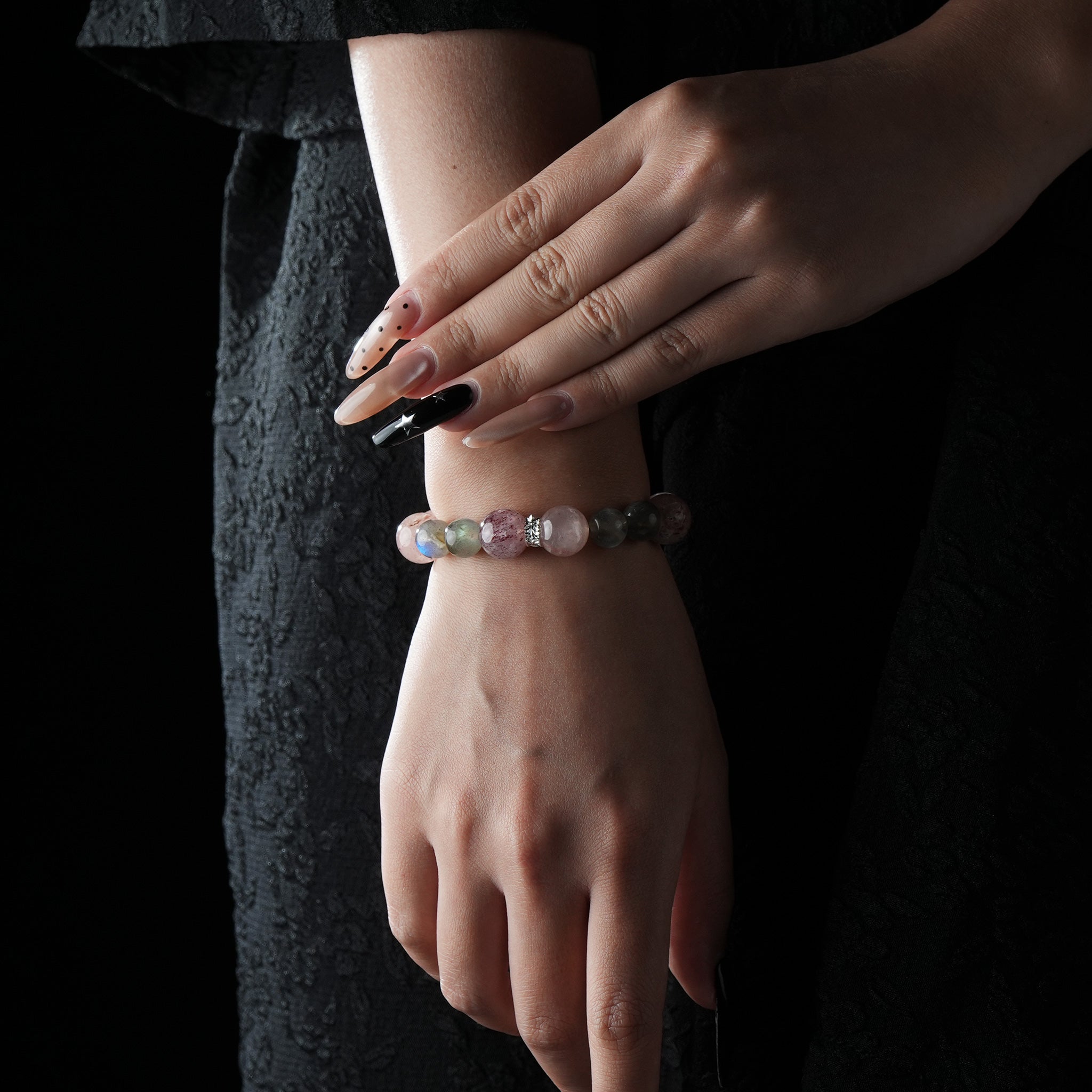 A model's wrist adorned with a soft-toned bracelet made of pink and gray beads, elegantly styled with dark clothing and long nails.