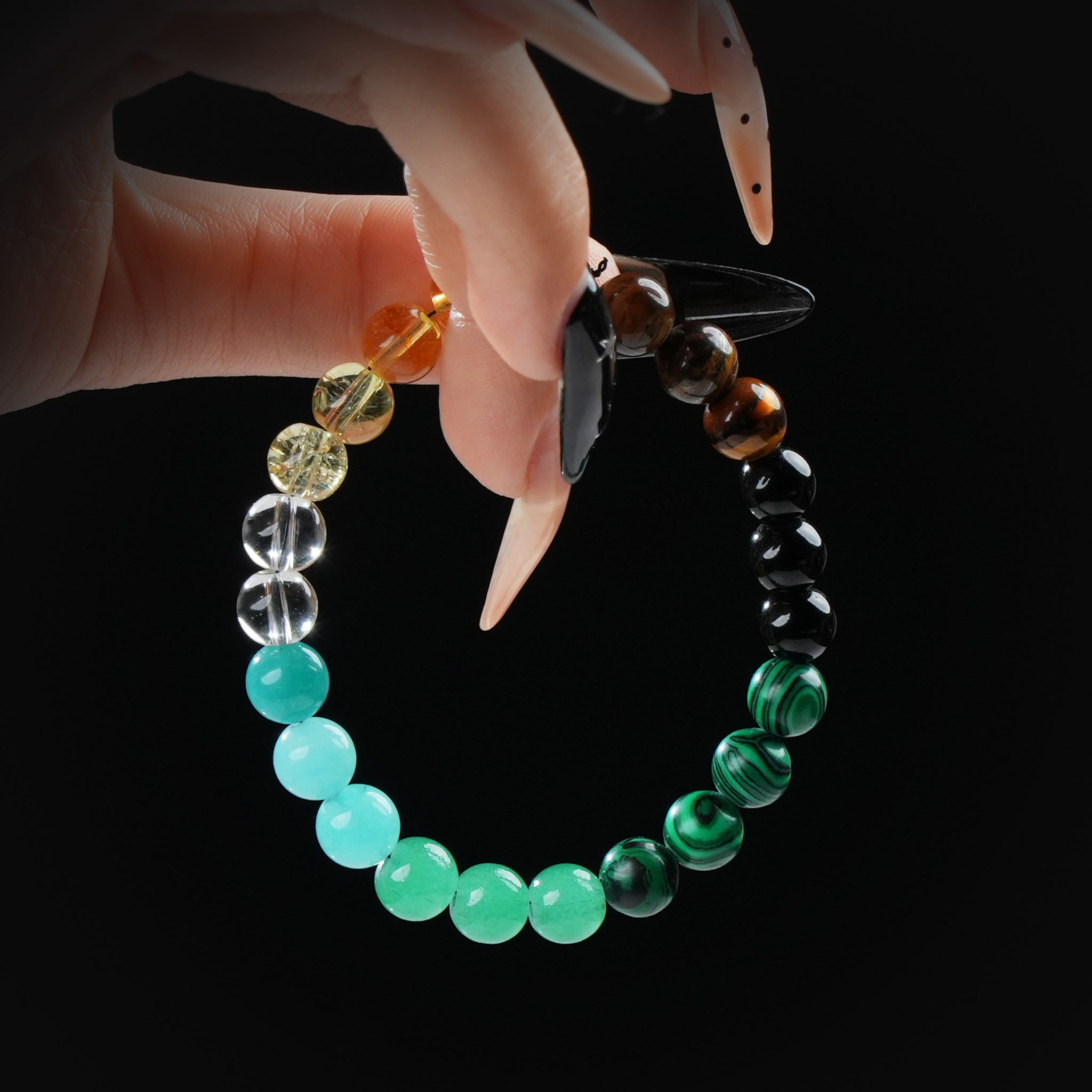 Bracelet composed of various gemstone beads in green, black, yellow, and brown shades, held by hand against a black background.