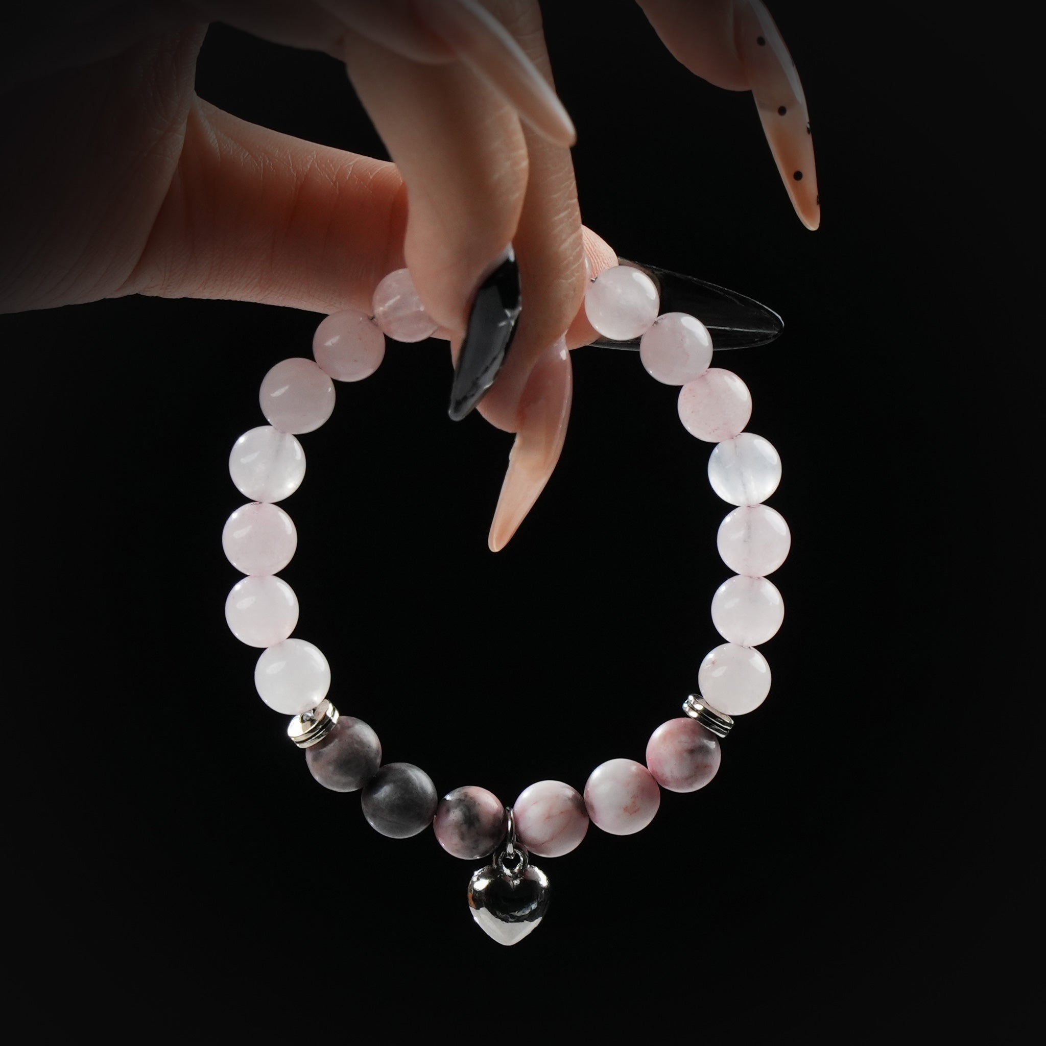 Delicate bracelet made of rose quartz and pink-toned beads, featuring a metallic heart charm, held by hand against a black background.