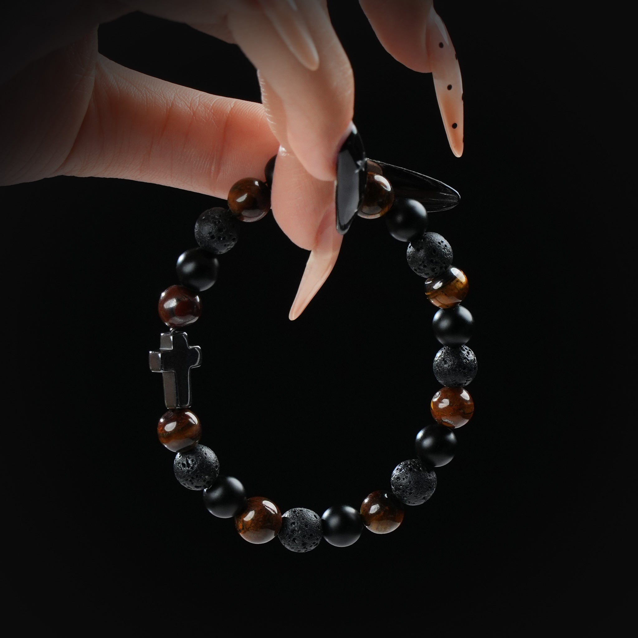 Tiger's eye, lava stone, and obsidian beaded bracelet with black cross charm, held by hand on black background for strength and faith.