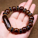 This image shows a close-up of a hand gently holding a 9-Eye Dzi bead bracelet. The centerpiece is a glossy, elongated Dzi bead with traditional Tibetan-style eye patterns, flanked by polished brown agate beads and black spacer rings bordered with gold accents. The overall design blends heritage symbolism with refined craftsmanship.