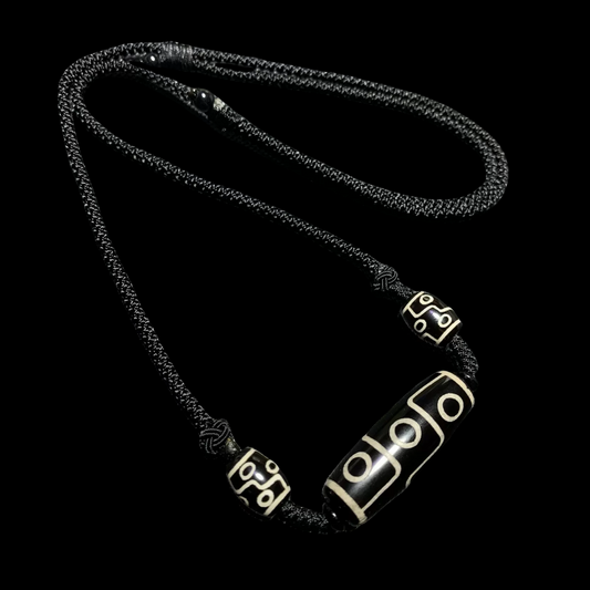 A close-up of a Tibetan Nine-Eyed Dzi bead necklace displayed on a textured black surface. The necklace features a central Dzi bead and two smaller beads, all on a black woven cord.