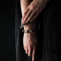 Elegant wrist shot of a woman wearing a beaded bracelet made of alternating clear quartz and gold sheen obsidian stones, styled with long manicured nails and a black textured dress.