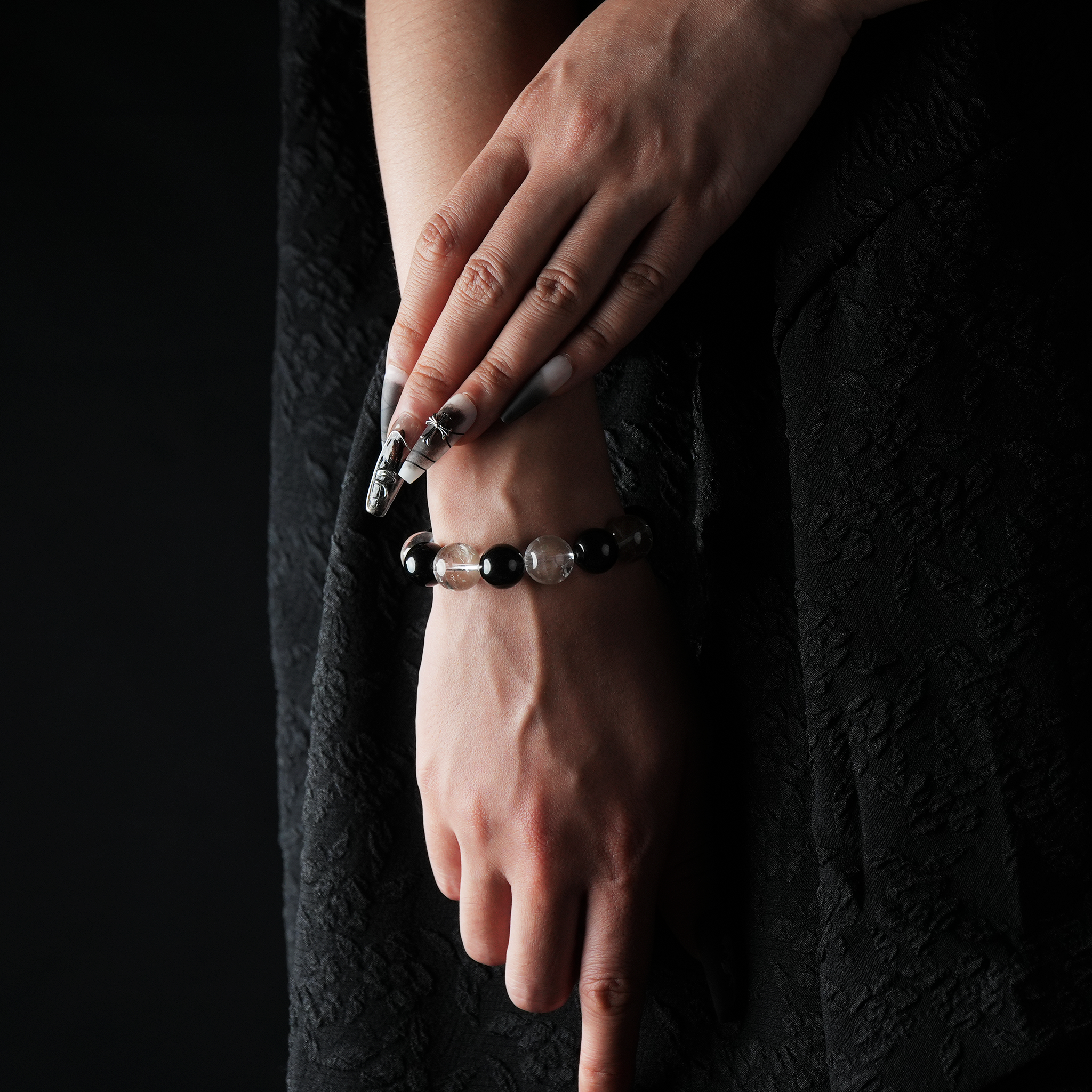 Elegant wrist shot of a woman wearing a beaded bracelet made of alternating clear quartz and gold sheen obsidian stones, styled with long manicured nails and a black textured dress.