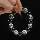 The image shows a hand holding a beaded bracelet composed of 12mm round Clear Quartz and Gold Sheen Obsidian gemstones. The Clear Quartz beads sparkle with clarity while the Gold Sheen Obsidian beads reflect subtle golden highlights, creating a striking contrast against the dark background.