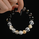 This photo captures a person gently holding a round gemstone bracelet between two fingers. The bracelet consists of polished 12mm beads in a gradient of tones: bright Clear Quartz, sunny Citrine, metallic Silver Obsidian, and dark glossy Obsidian. The stones’ textures and reflections are emphasized by the clean black backdrop and soft lighting.