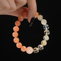 In this image, a hand delicately holds a round beaded bracelet made of natural gemstones. The bracelet features soft orange moonstone beads on one side and bright yellow citrine on the other, with a central bead of red rutilated quartz standing out for its internal coppery threads. The black background adds contrast to highlight the warm colors of the stones.